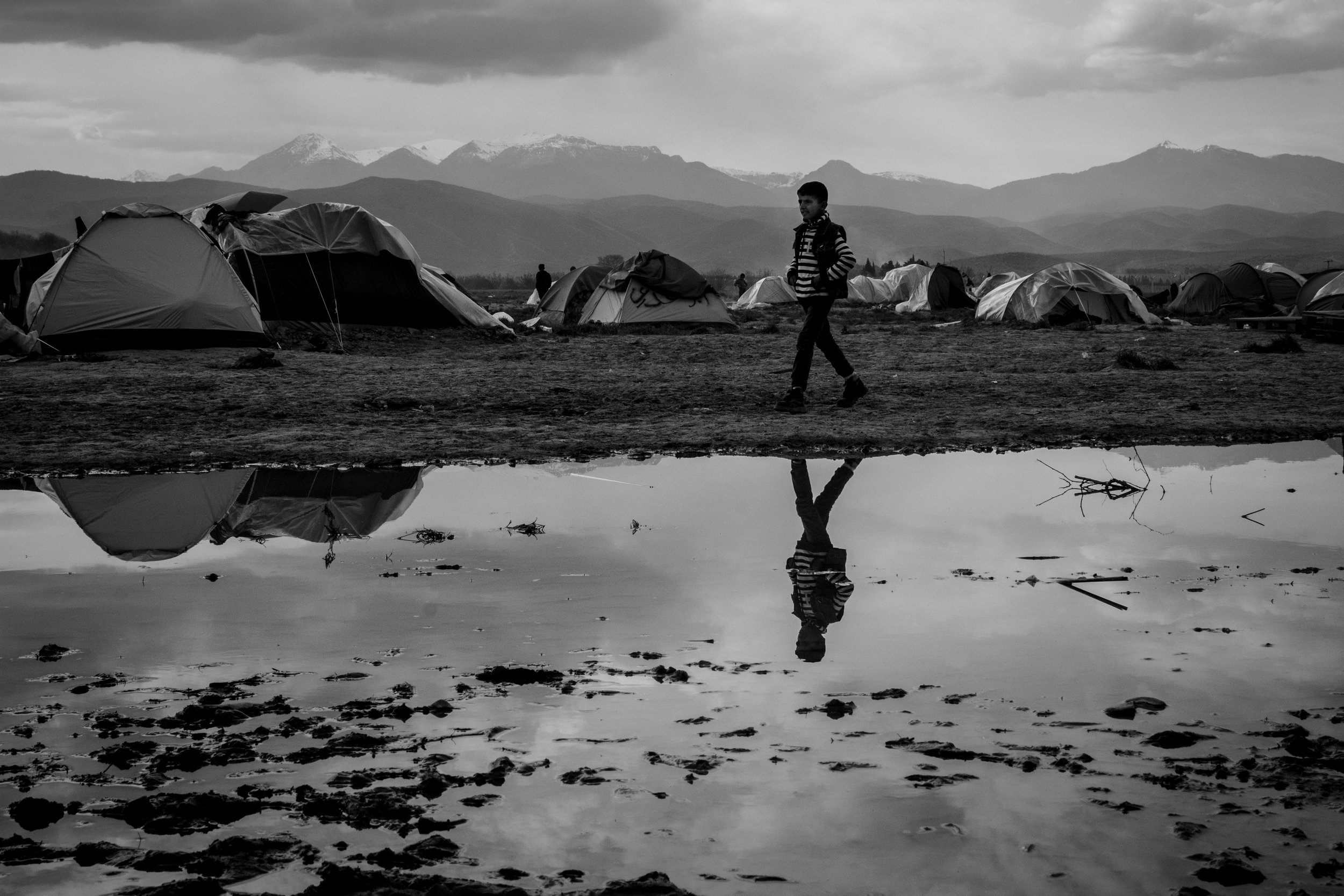 A boy walks from his family's tent into the centre of the camp, with Macedonian mountains behind him; they represent the European haven that lies beyond the border.