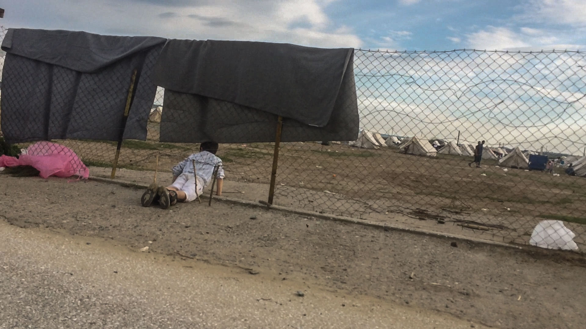 A refugee boy crawls in and out of the low wire fencing surrounding Nea Kavala camp. This was ironic because a hundred metres or so along the fence were numbers of police guards and dogs.