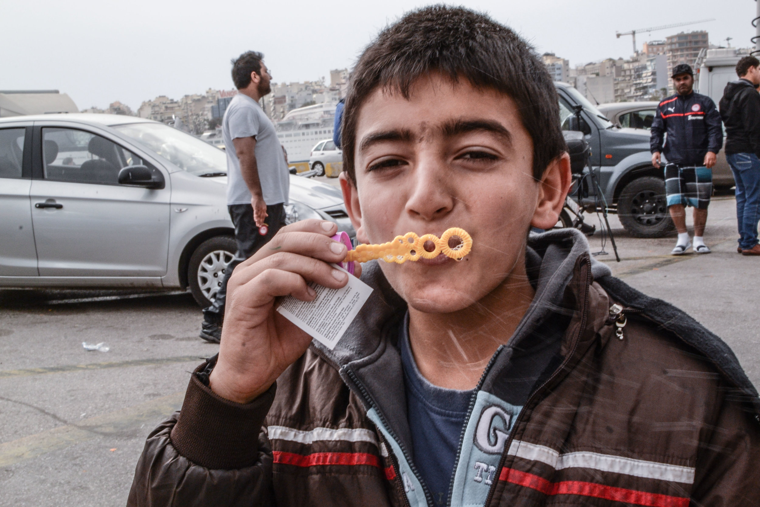 A young refugee happily blows bubbles near his family’s tent in Piraeus port, in spite of the tumultuous crowd waiting for medical supplies from the Greek Red Cross behind him.
