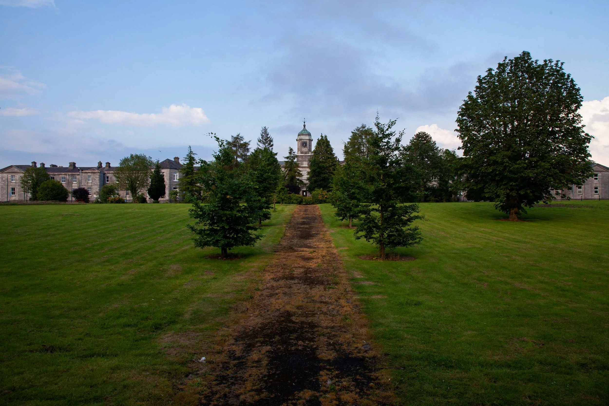   St. Fintan's Hospital, Portlaoise (previously Maryborough District Lunatic Asylum)   Est: 1833 – Currently in operation (partially)  Front exterior with main entrance (shrouded by trees) and pathway leading to formal gardens and summer house pavili