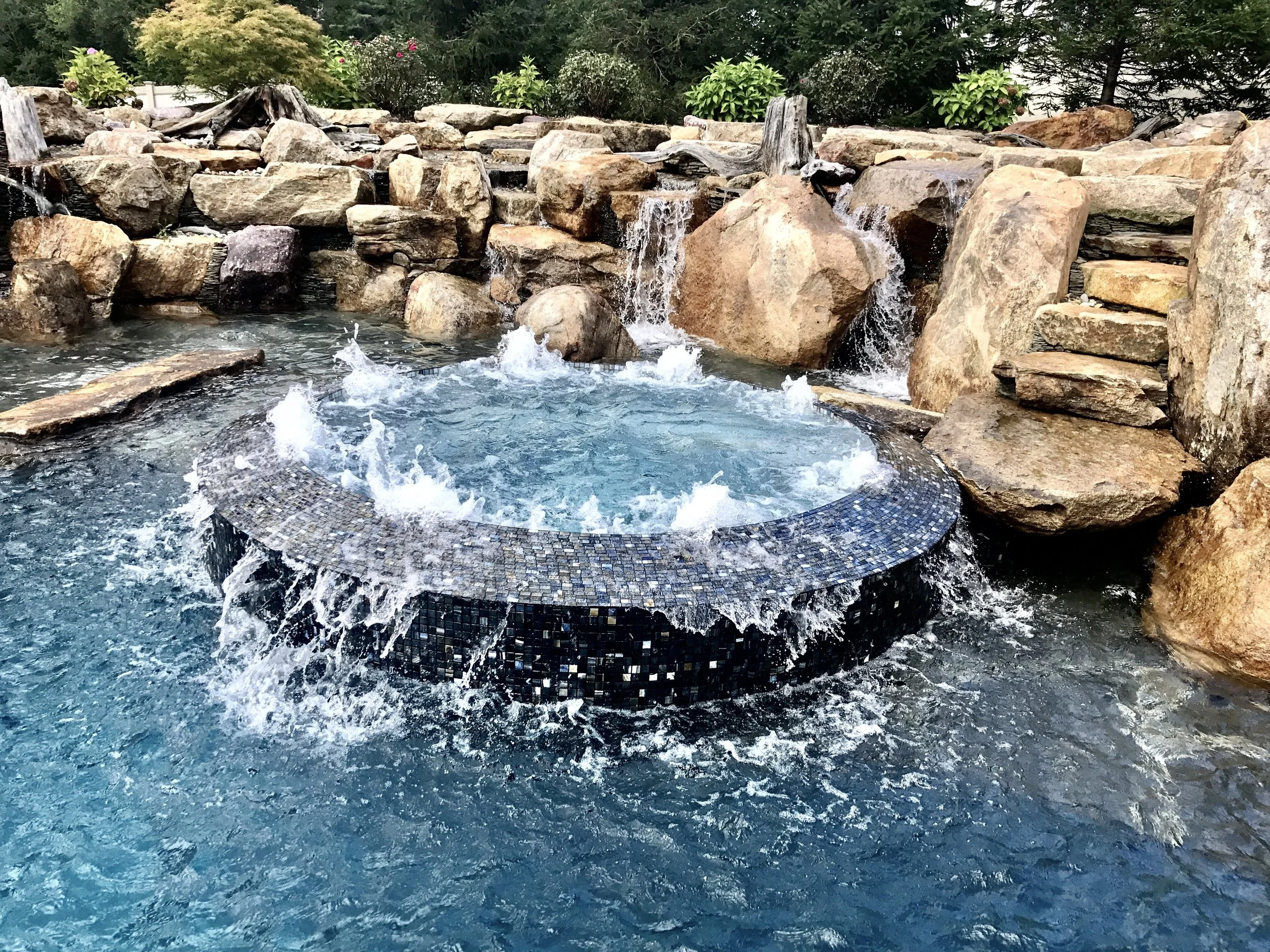 A diving rock and spa are set atop a deep end swim shelf. Glass mosaic tile covers the spa and spa spillway.