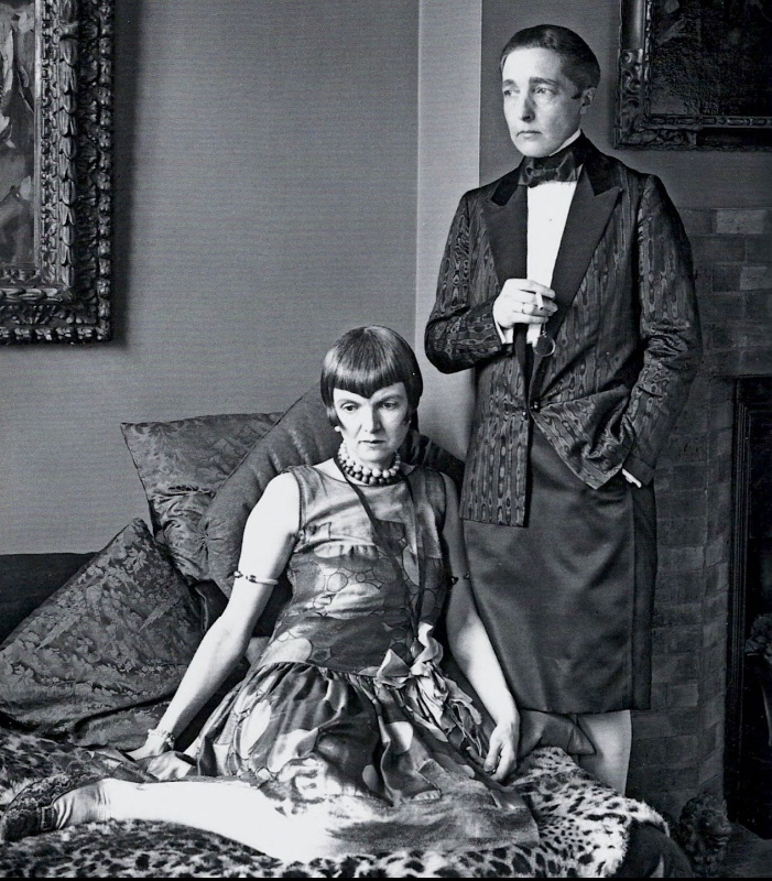 Radclyffe Hall standing wearing a pencil skirt, blazer, bowtie, and holding a monocle next to Una Troubridge wearing a dress and various accessories.