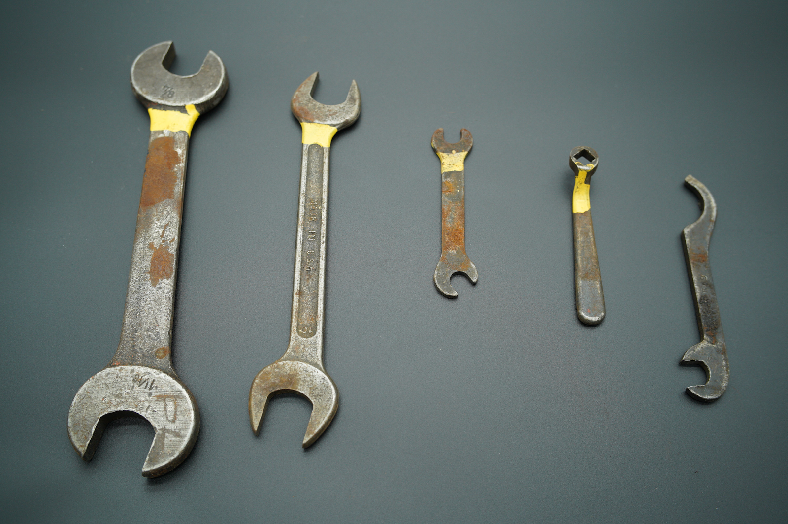 Photograph of five rusty wrenches arranged in a row on a dark surface, varying in size from large to small. Each wrench has a yellow tape mark near the head, highlighting wear and corrosion on the metal.