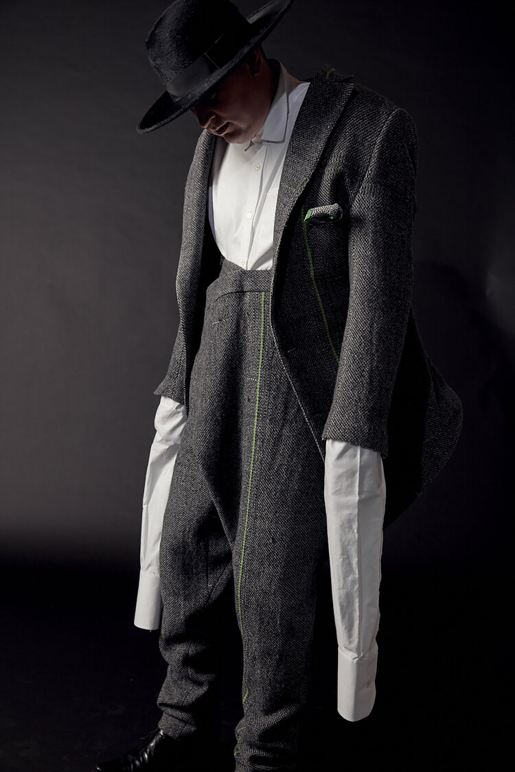 Philip, head downcast and covered in shadow from his hat. His shirt sleeves spill out from his jacket, and his trousers drape along the center front before bunching around his shoes.