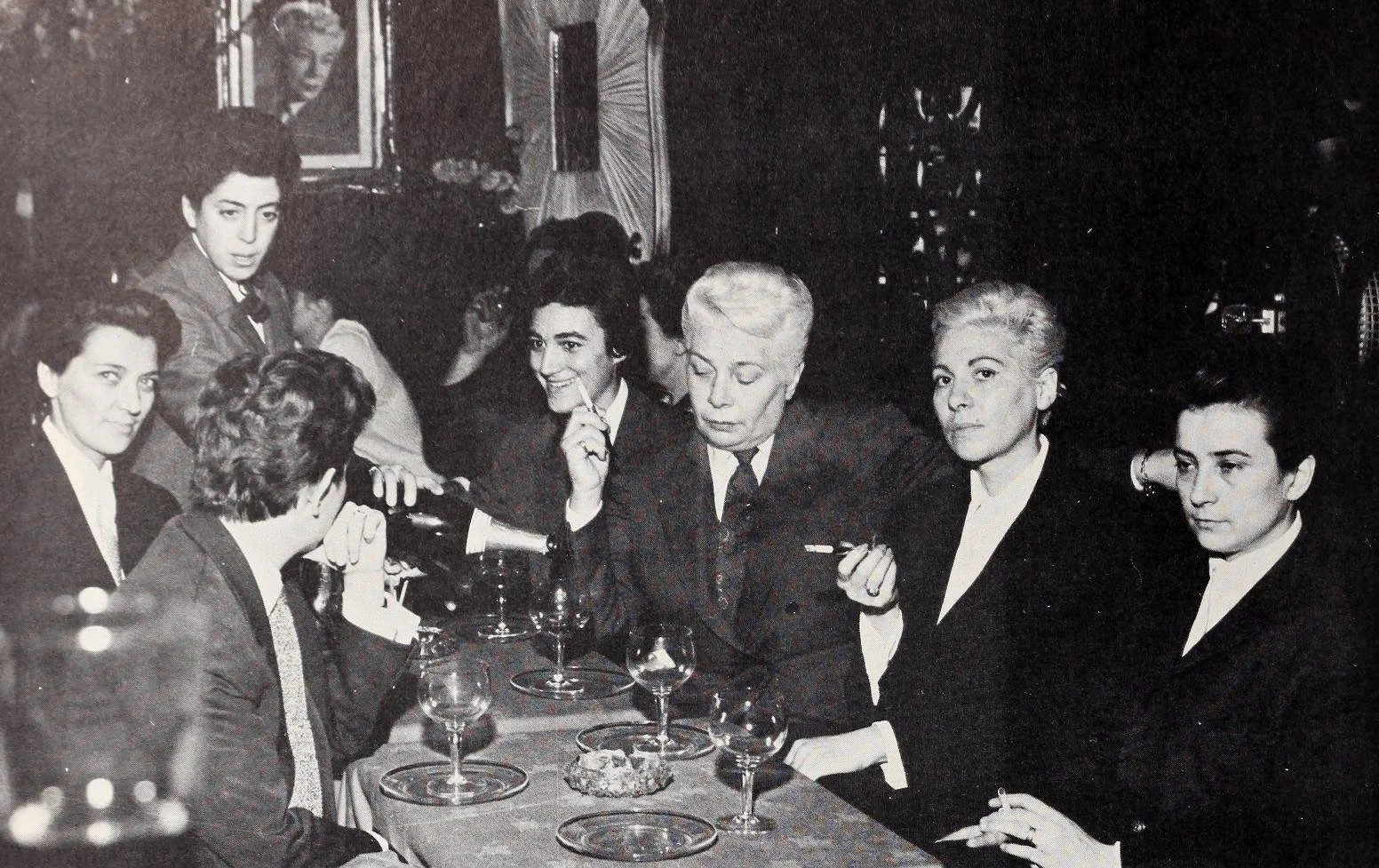 Moune wearing a tie and blazer and smoking a cigarette at a table with various guests of the bar Chez Moune dressed in a similar style.