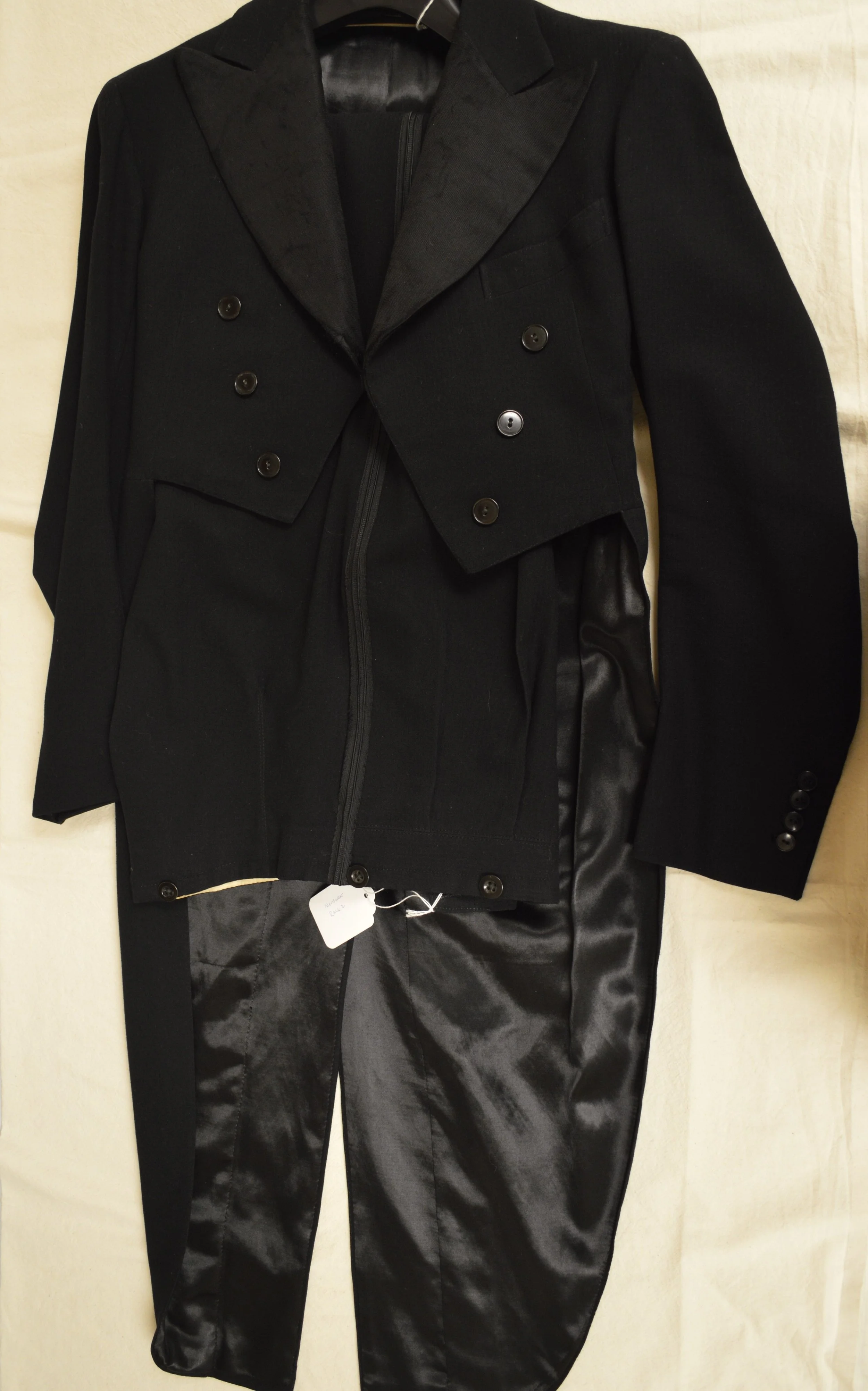 The FRC tailcoat is laid flat on a table with its arms at its sides. The corresponding pants are folded on the hanger beneath them, the archival label just slightly visible.