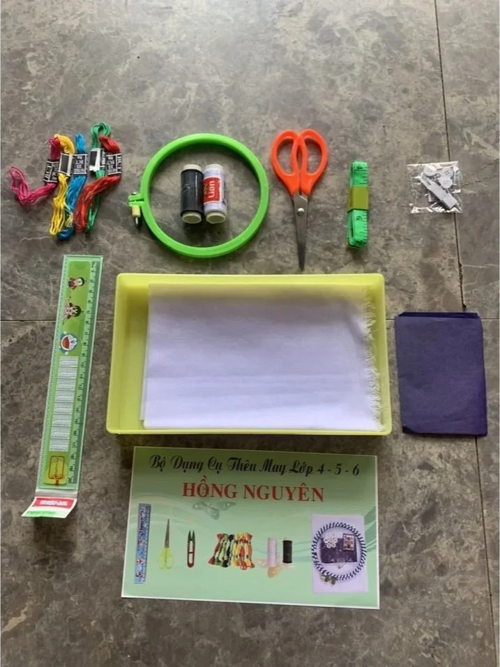 Complimentary embroidery kit for “Kĩ-Thuật” laid out on a grey laminate floor. The kit includes a hoop, a piece of fabric, scissors, threads, tracing paper, rulers, and buttons.
