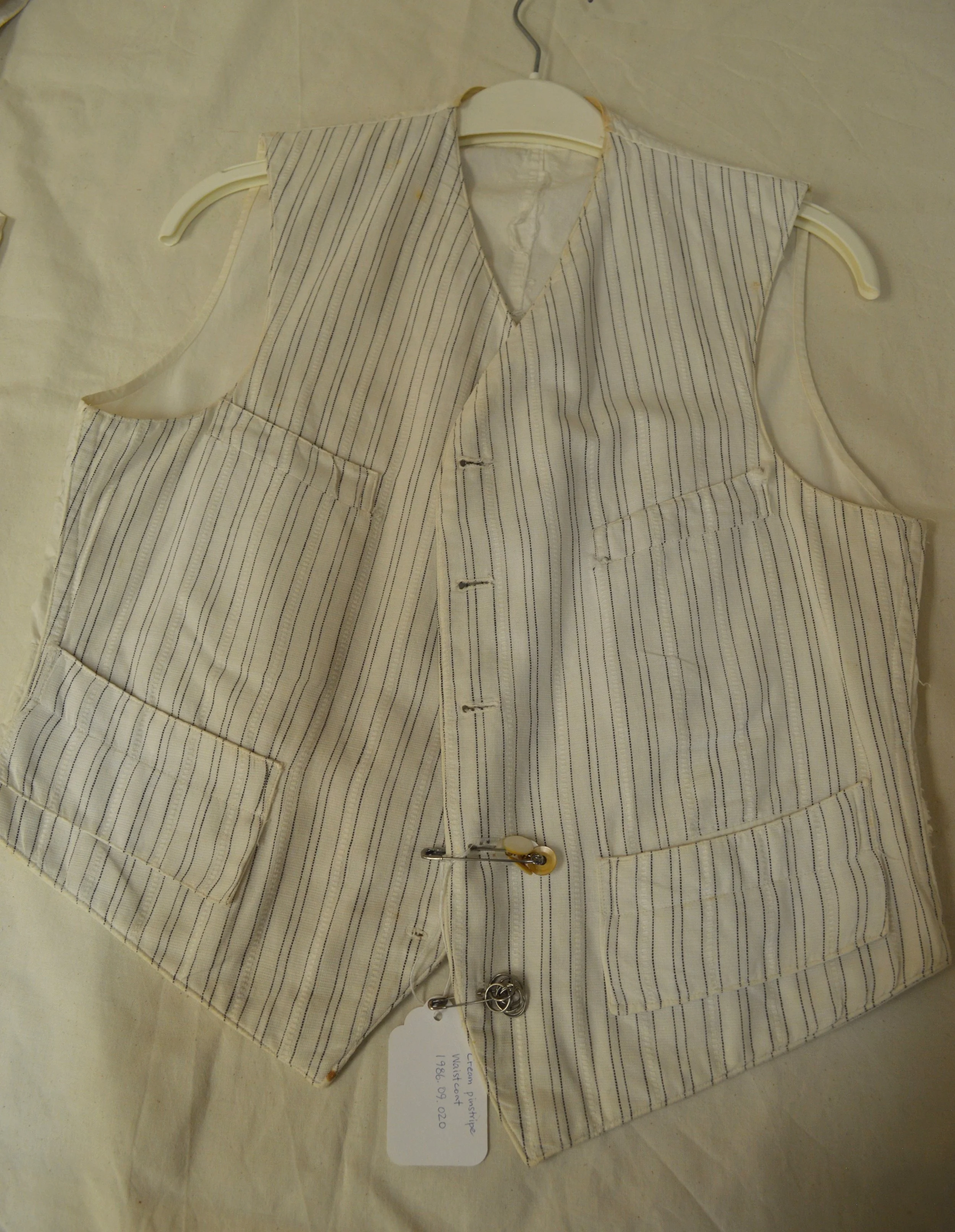 A top-down view of a white cotton vest laid flat on a table covered in archival paper. The vest has black pinstripes, detached pearl buttons held by safety pin to its front panel, and four pockets.