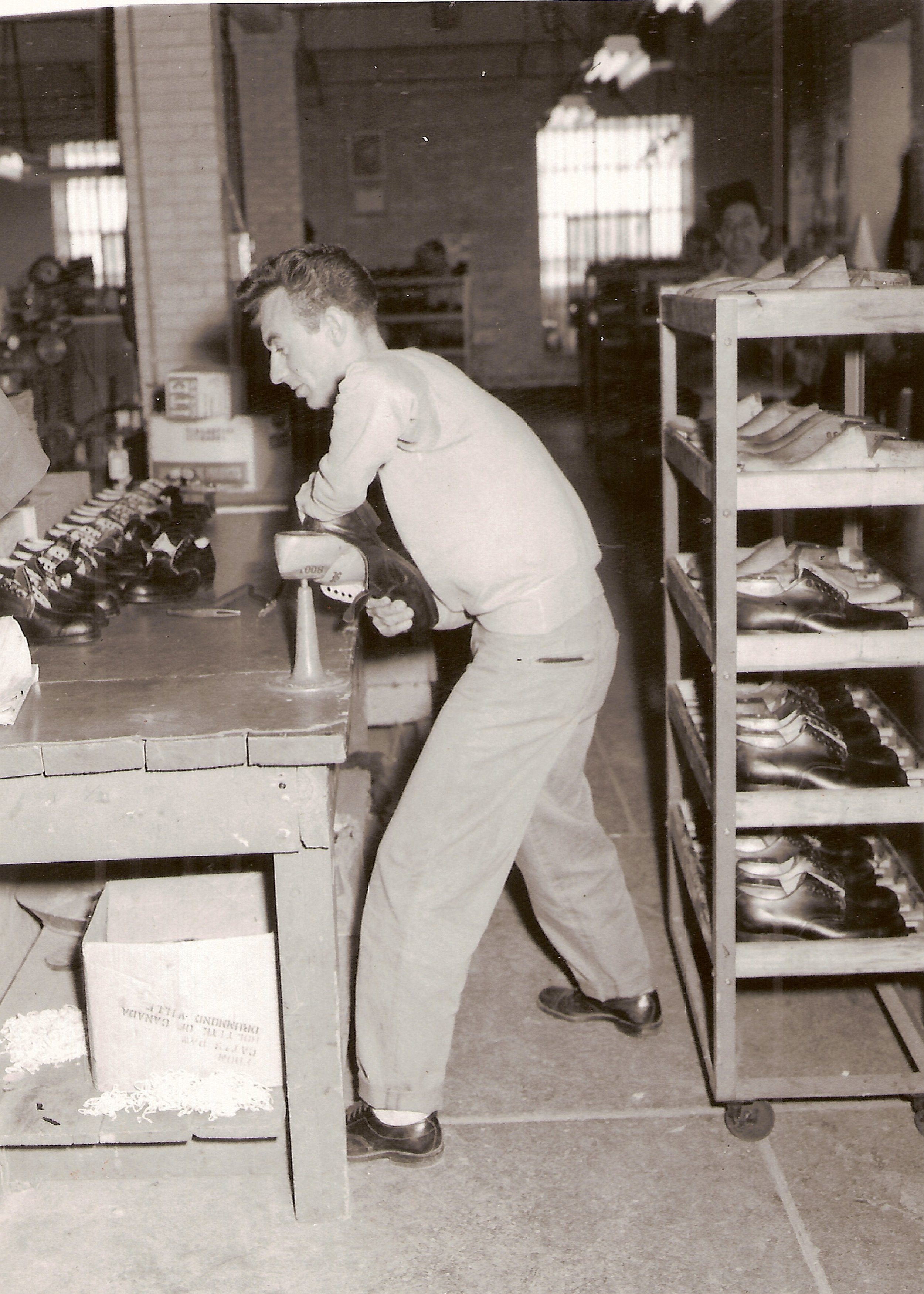 Man working in a shoe workshop, standing at a workbench with various tools and shoes. Shelves filled with multiple pairs of shoes are visible to the right, indicating an organized workspace focused on shoe production.