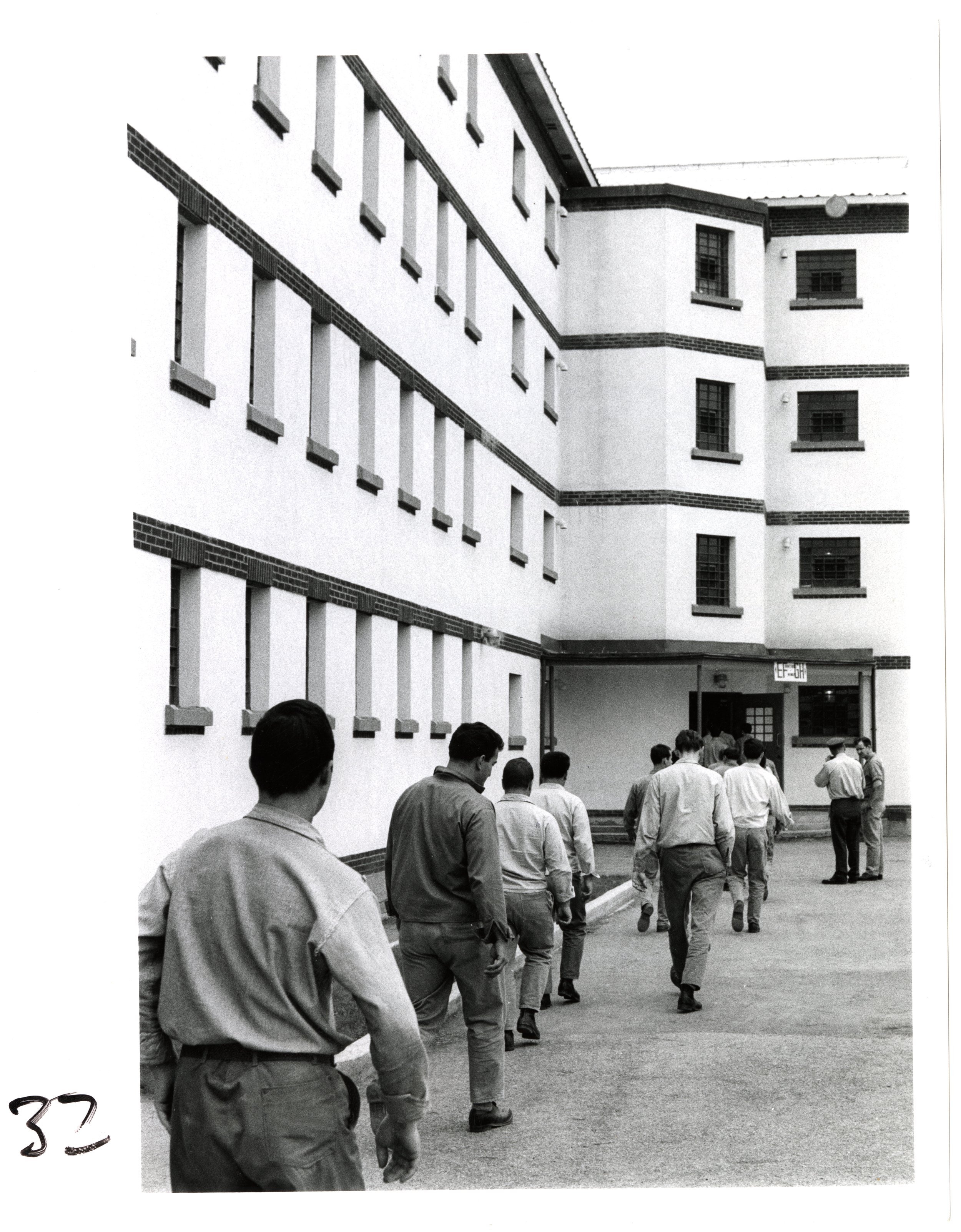 A group of men walking in a line toward a prison building with multiple windows and a sign above the entrance. The men are dressed in similar work style clothing and walking in an orderly manner.