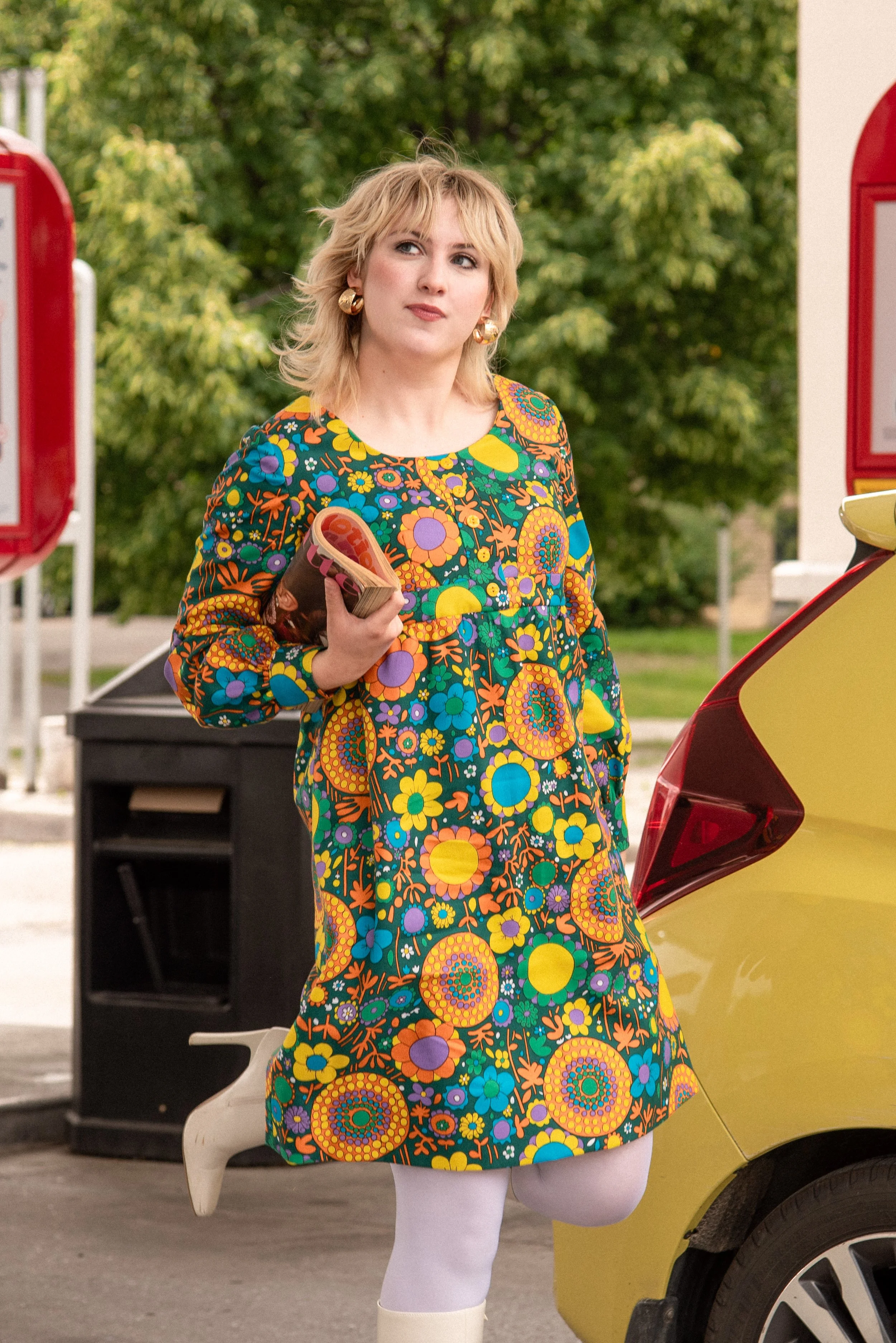 Designer Natalie Welsh skipping across a gas station holding a magazine wearing a bright floral patterned cotton dress and white gogo boots.