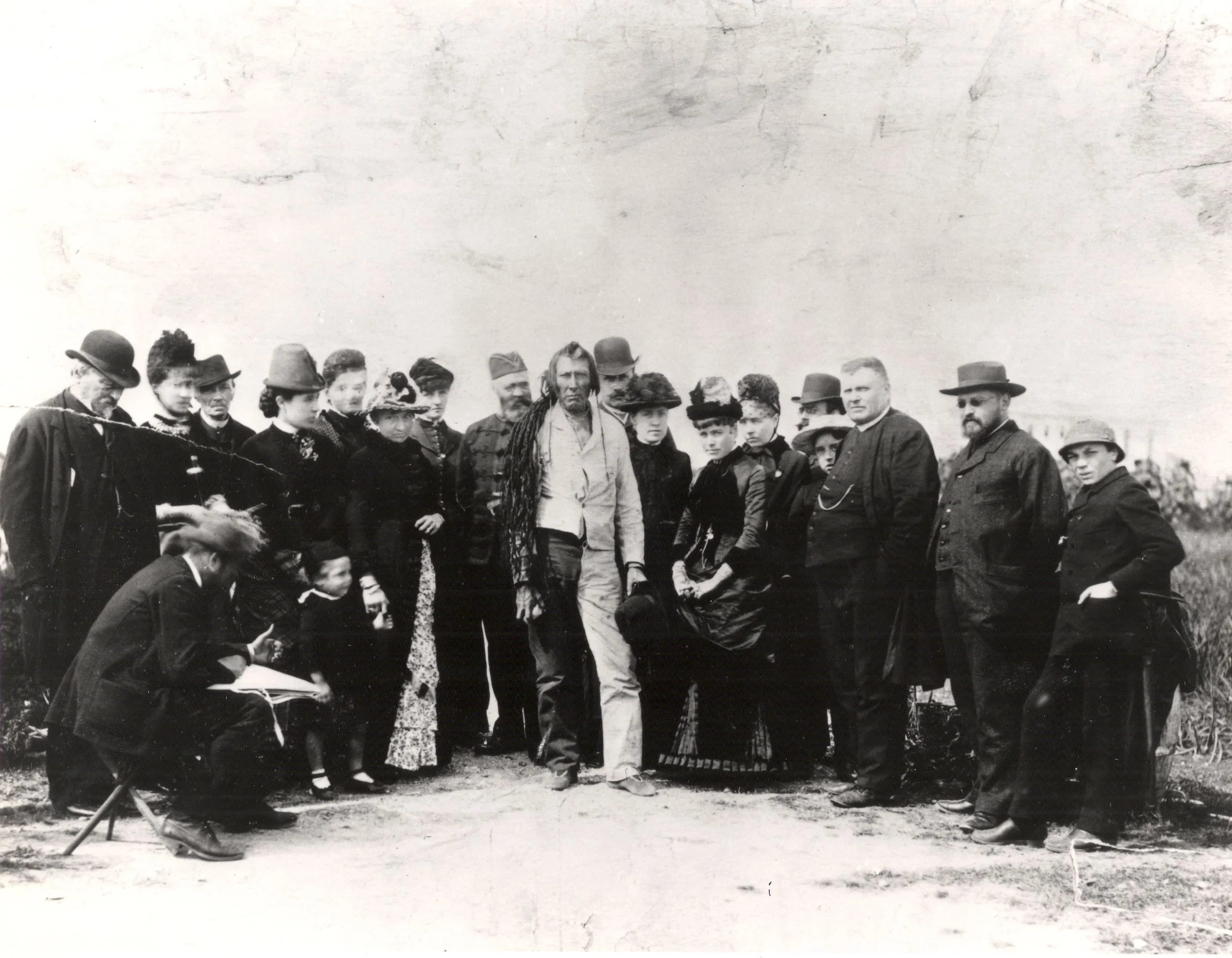 Group of men and women dressed in late 19th-century attire gathered outdoors around a central Indigenous man. The scene captures a moment of interaction and also documentation, with one man seated and writing in a notebook.