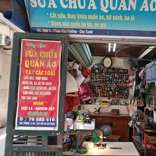 A storefront of a mending service shop in Hanoi, Vietnam. Behind the small desk is a small fan, lamp, and a variety of fabric and mending tools hanging on the walls.