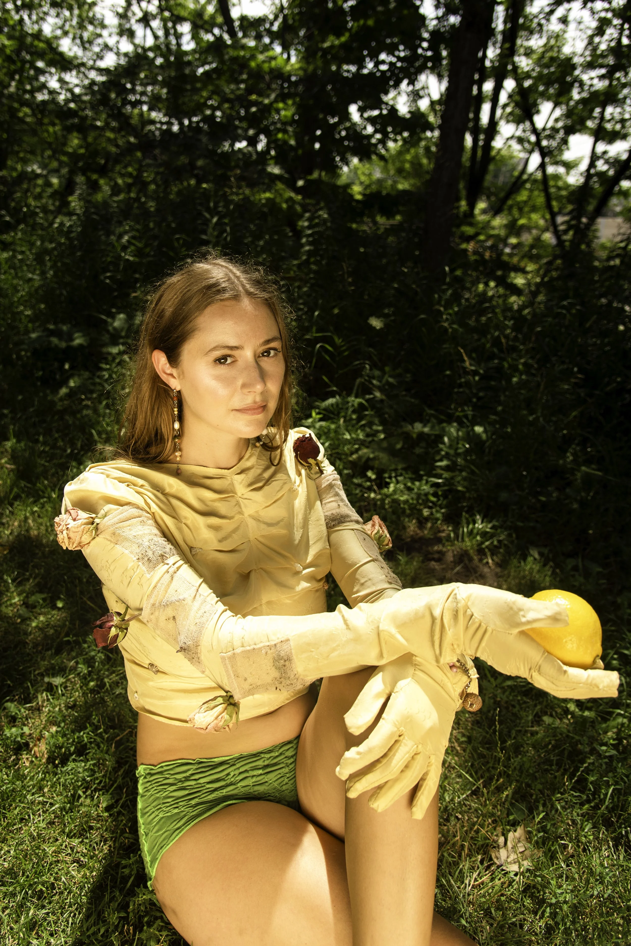 Designer Isla Cowan seated in a sunlit forest, wearing a sculptural yellow long sleeve silk top with attached gloves and floral details, holding a lemon.
