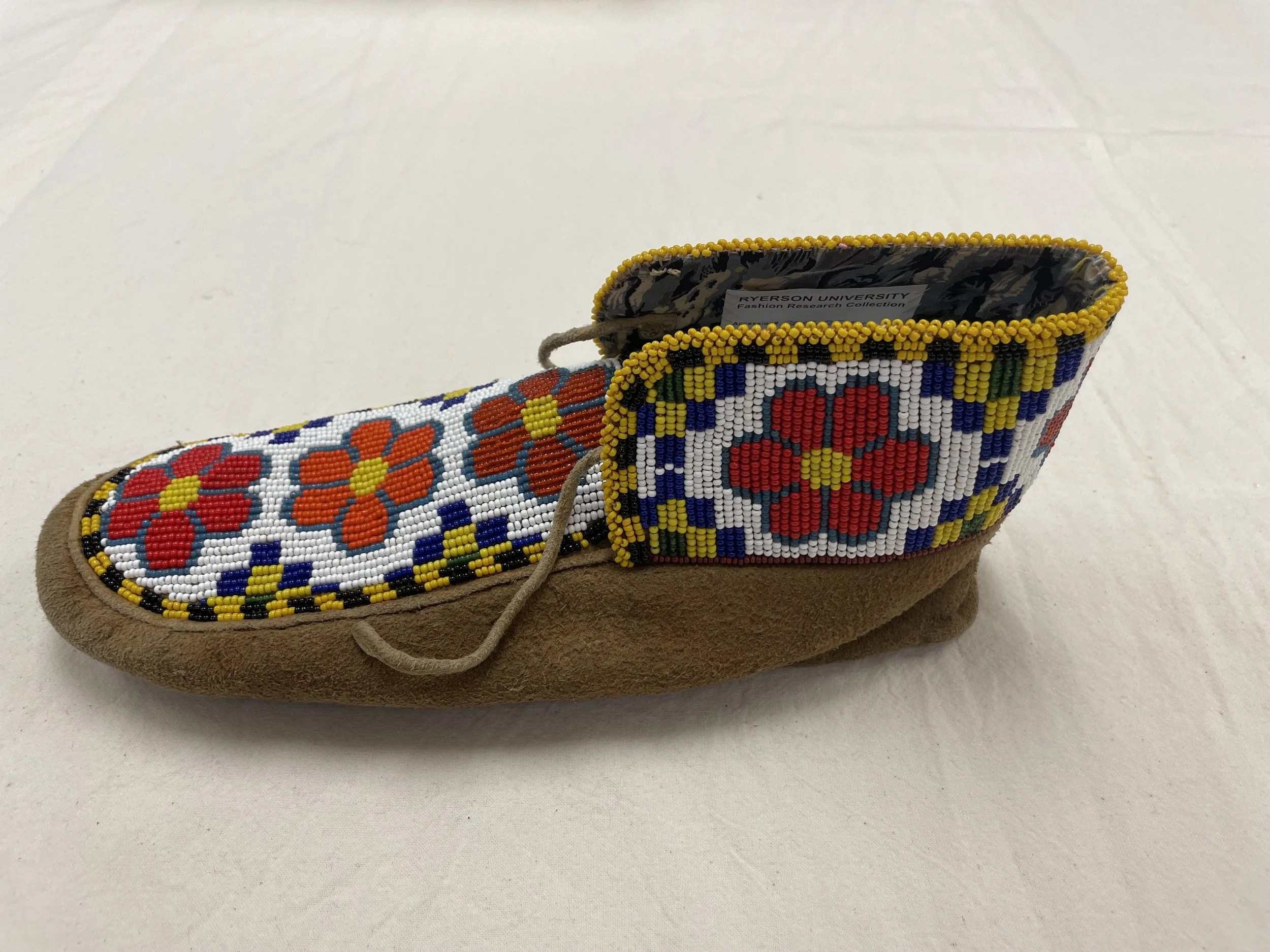 Side profile of a moccasin featuring intricate beadwork with red and orange flowers, blue and yellow geometric patterns, and a yellow border on a tan suede base. The moccasin has a lace closure and a soft sole.