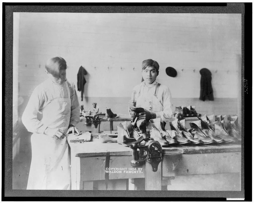 Two young adolescent men in aprons standing at a workbench filled with various shoes and shoe repair tools. The setting is a shoe workshop at a residential school.