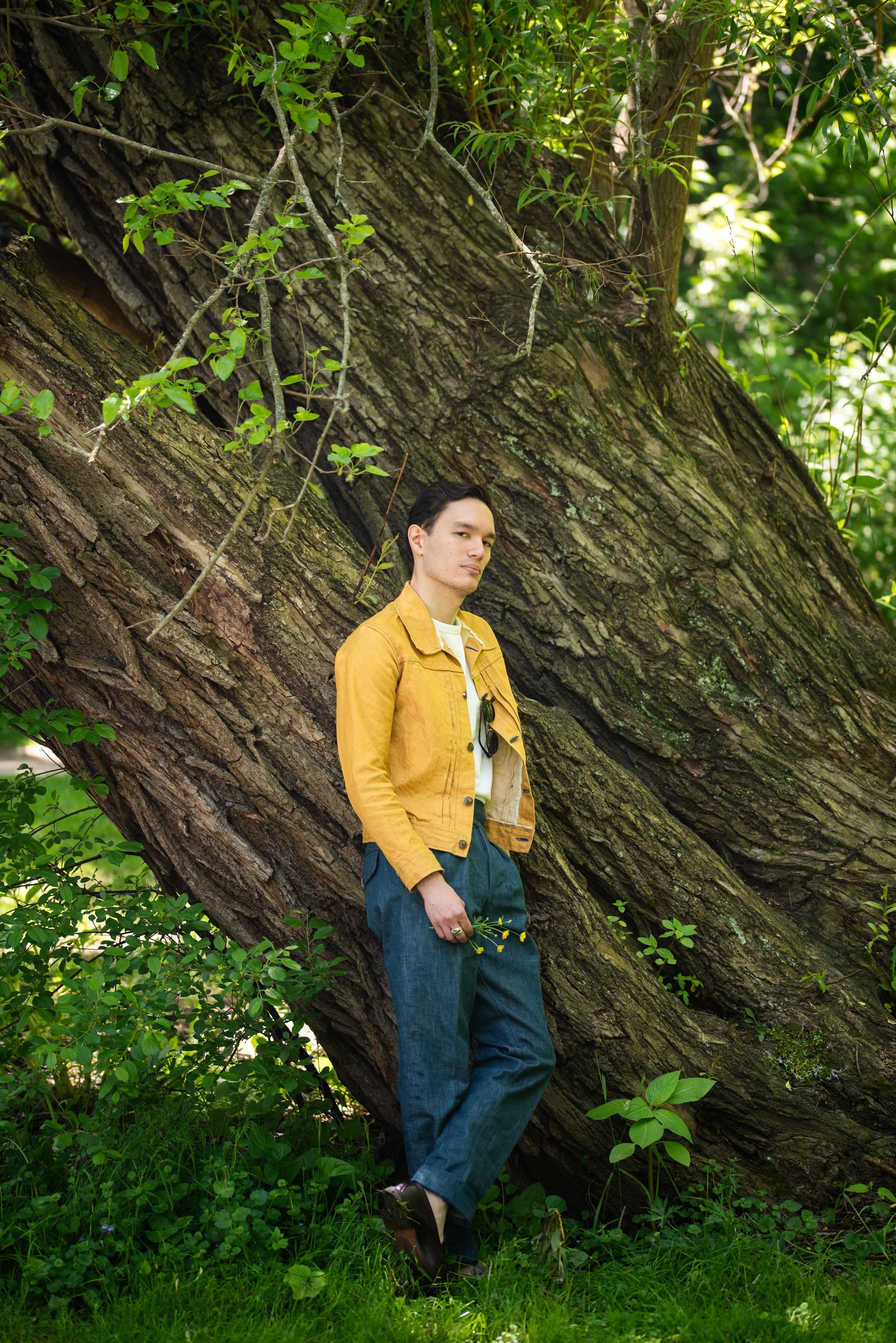 Designer Eric Larsen standing and leaning against a large tree with one leg crossed over the other while holding buttercup flowers, wearing a yellow denim jacket, white shirt and blue denim pants.
