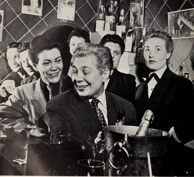 Moune, owner of Chez Moune, wearing a patterned tie and blazer sitting at the bar of the establishment with other butch presenting figures and waiters behind them.