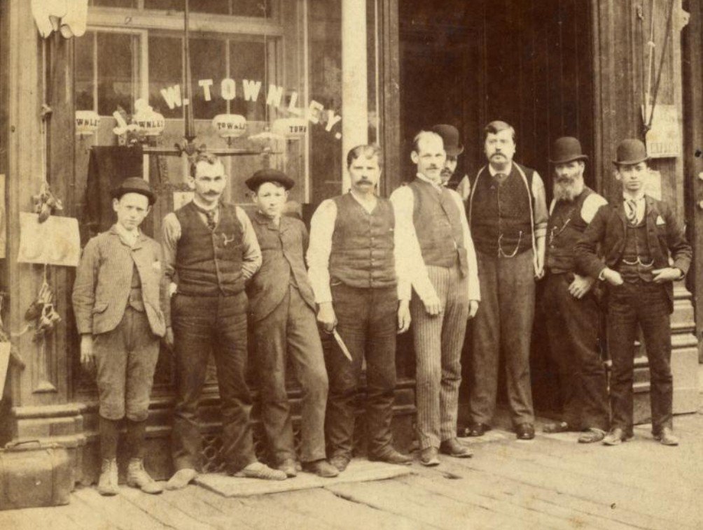 Nine male tailors of mixed ages stand in front of their shop, where J. & W. Townley is printed on the window. The photograph is sepia-toned.