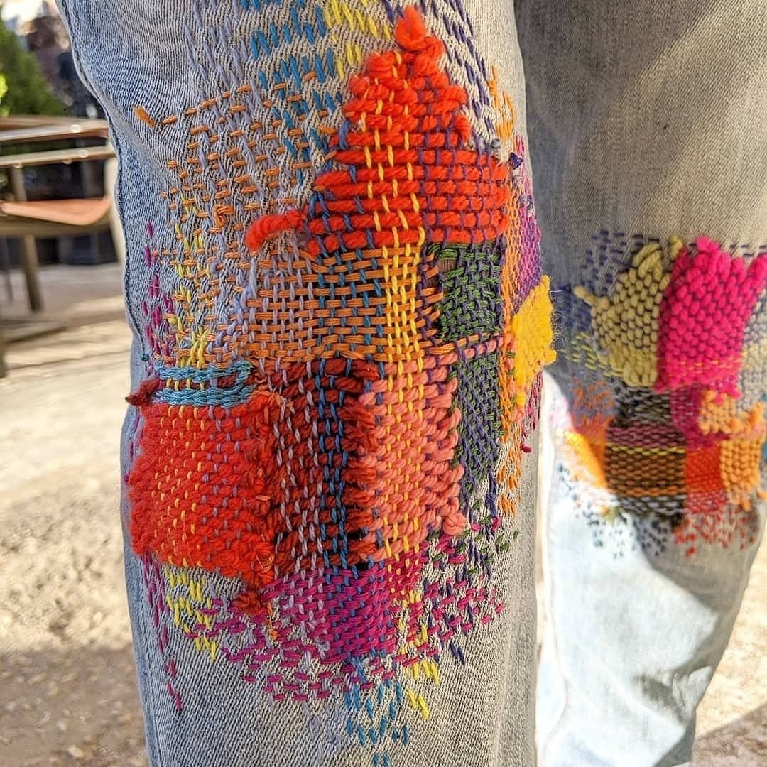 Sashiko mending involving many high-chroma coloured stitches woven on the knee of a pair of light wash jeans.