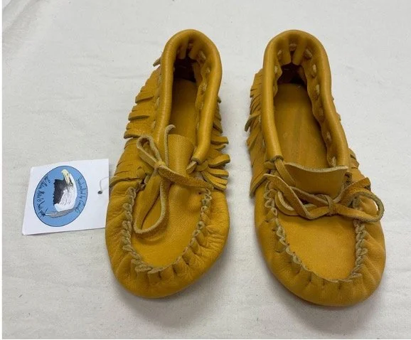 Pair of leather moccasins with leather fringe detailing along the sides and leather laces tied in bows on top. Shoes are placed on a white fabric surface, with the sales tag featuring a circular logo with a bird illustration.