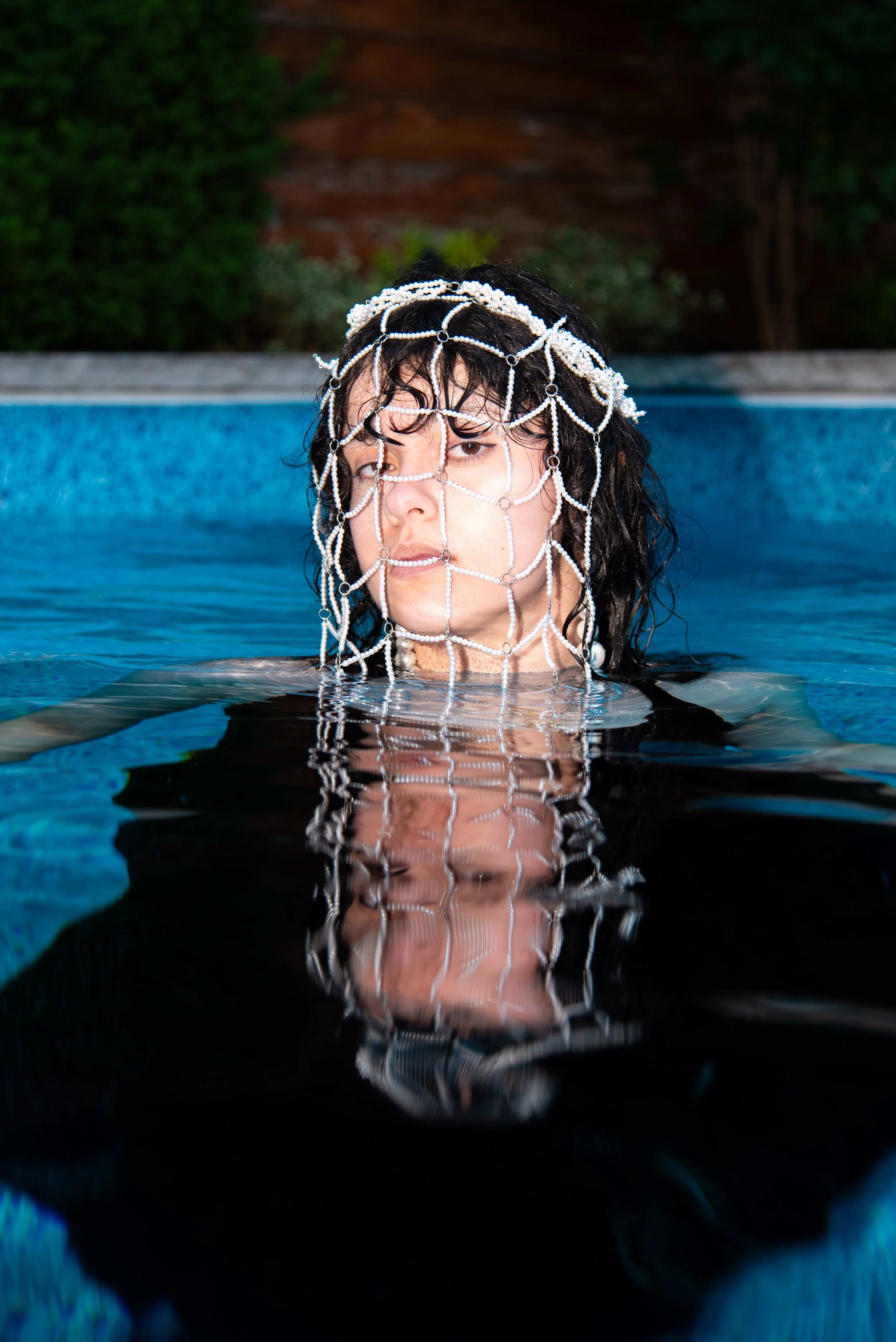 Designer Delfina Russo submerged in a pool up to her neck, wearing pearl and chainmail jewelry over her face.