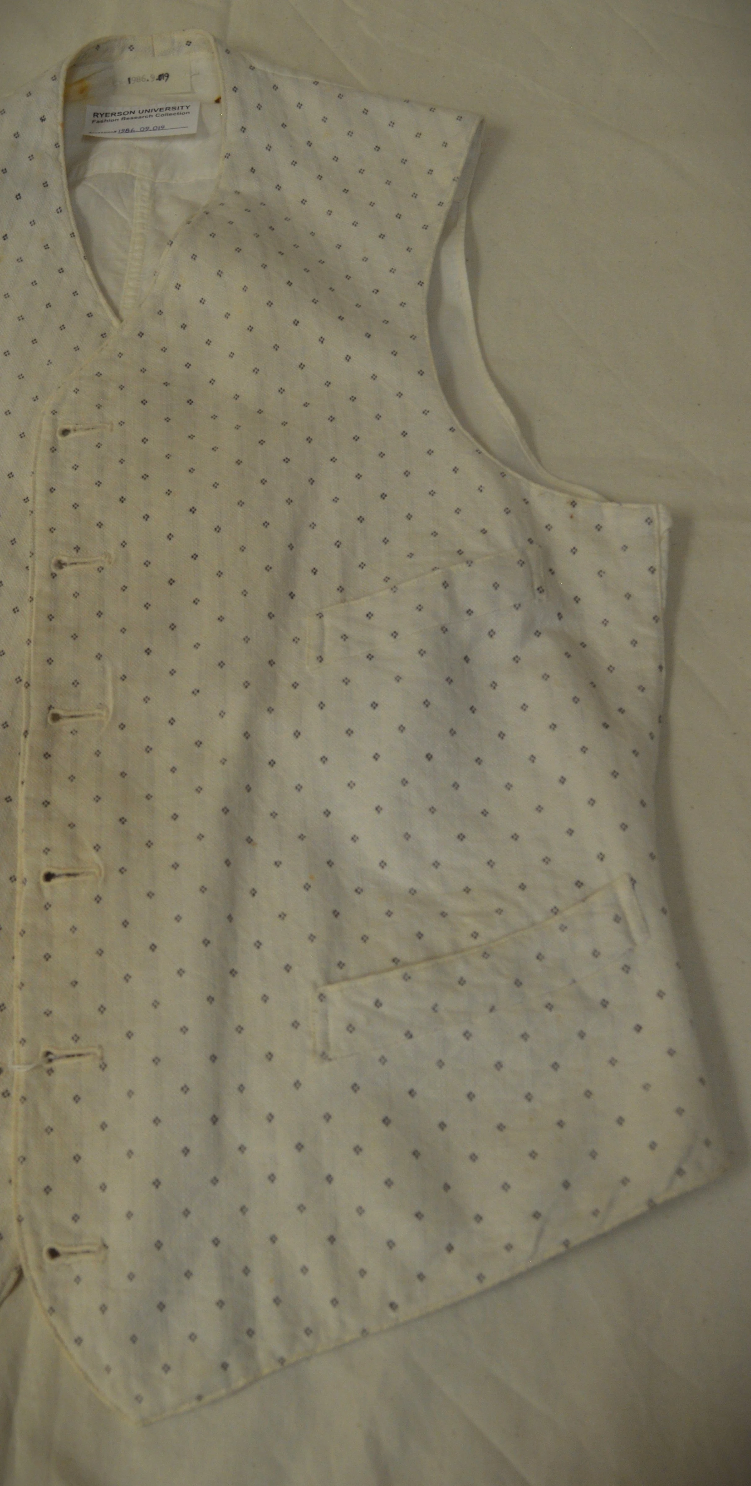 The left side (when worn) of a white cotton vest with black dots from the FRC. The vest is laid flat on archival paper.