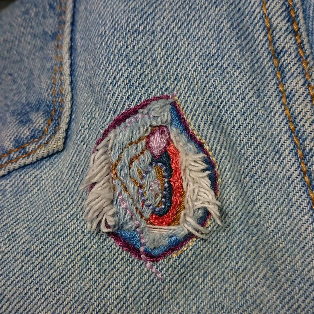Visible mending on a pair of blue jeans using a variety of bright colours. The patch is in the shape of a vulva.