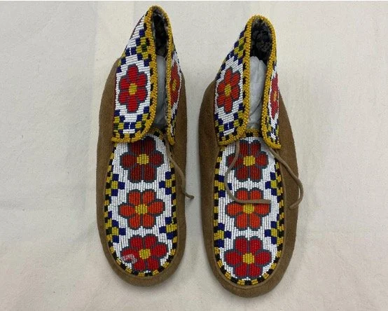Pair of moccasins with intricate beadwork on the top. Beadwork features red and orange flowers with yellow and blue geometric patterns on a white background, contrasting with brown suede sides.