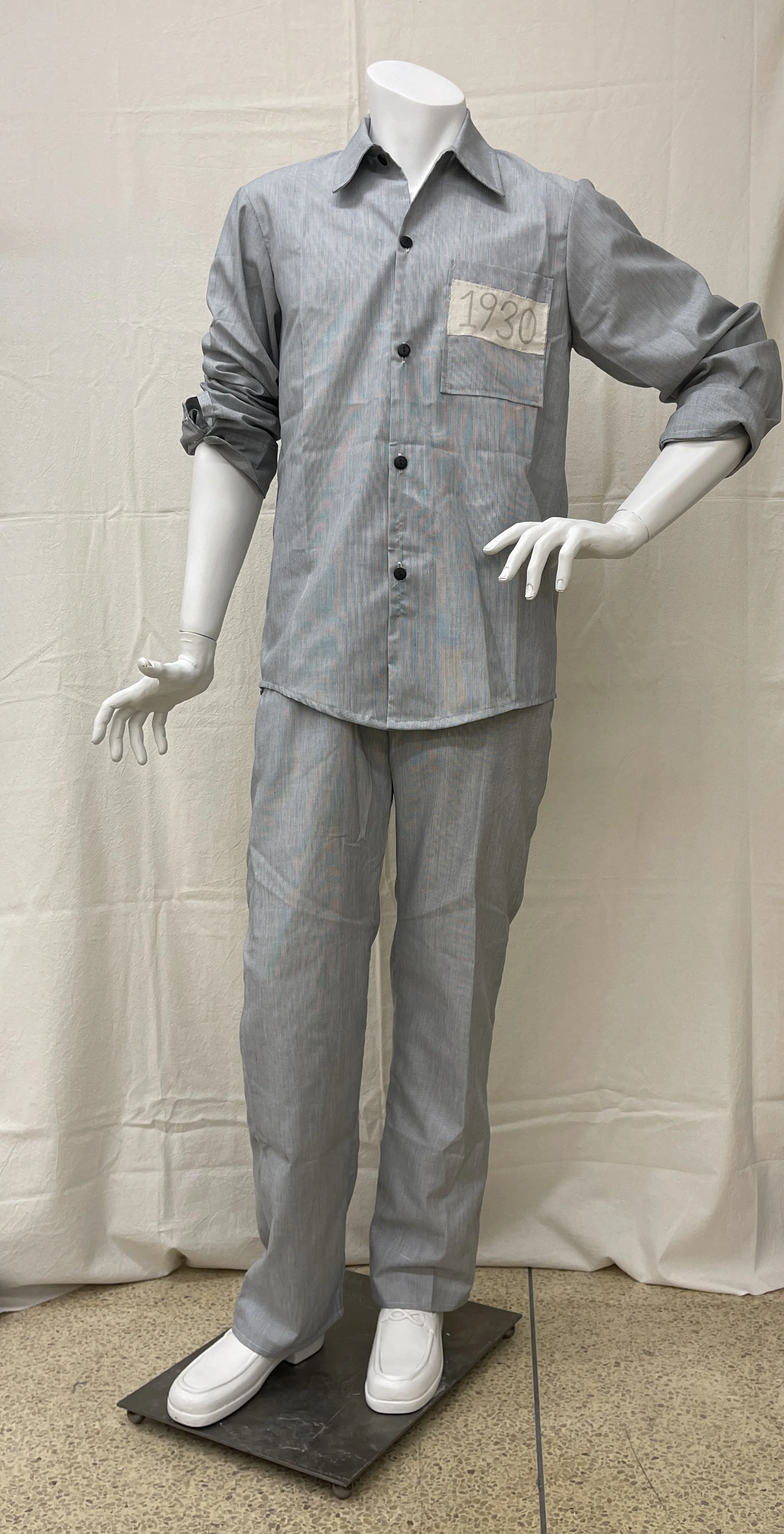 Photograph of a mannequin dressed in a grey striped prison uniform with a patch labeled "1930" on the chest. The outfit includes a long-sleeve button-up shirt with rolled sleeves, matching pants, and white slip-on shoes.