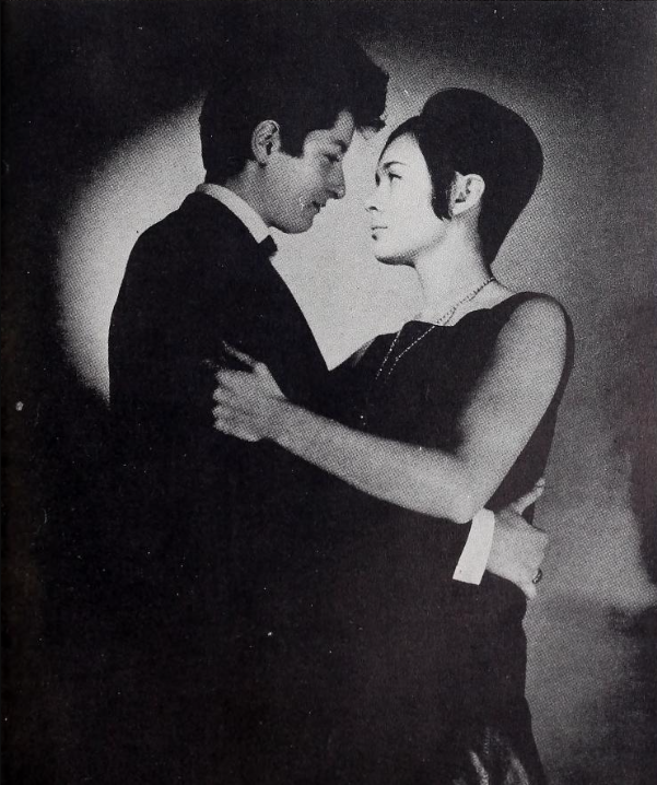 Male impersonator Marie Pierre wearing a suit and sporting a cropped haircut holding a more femme presenting woman in a dress and in a more typical 1960s women's haircut.