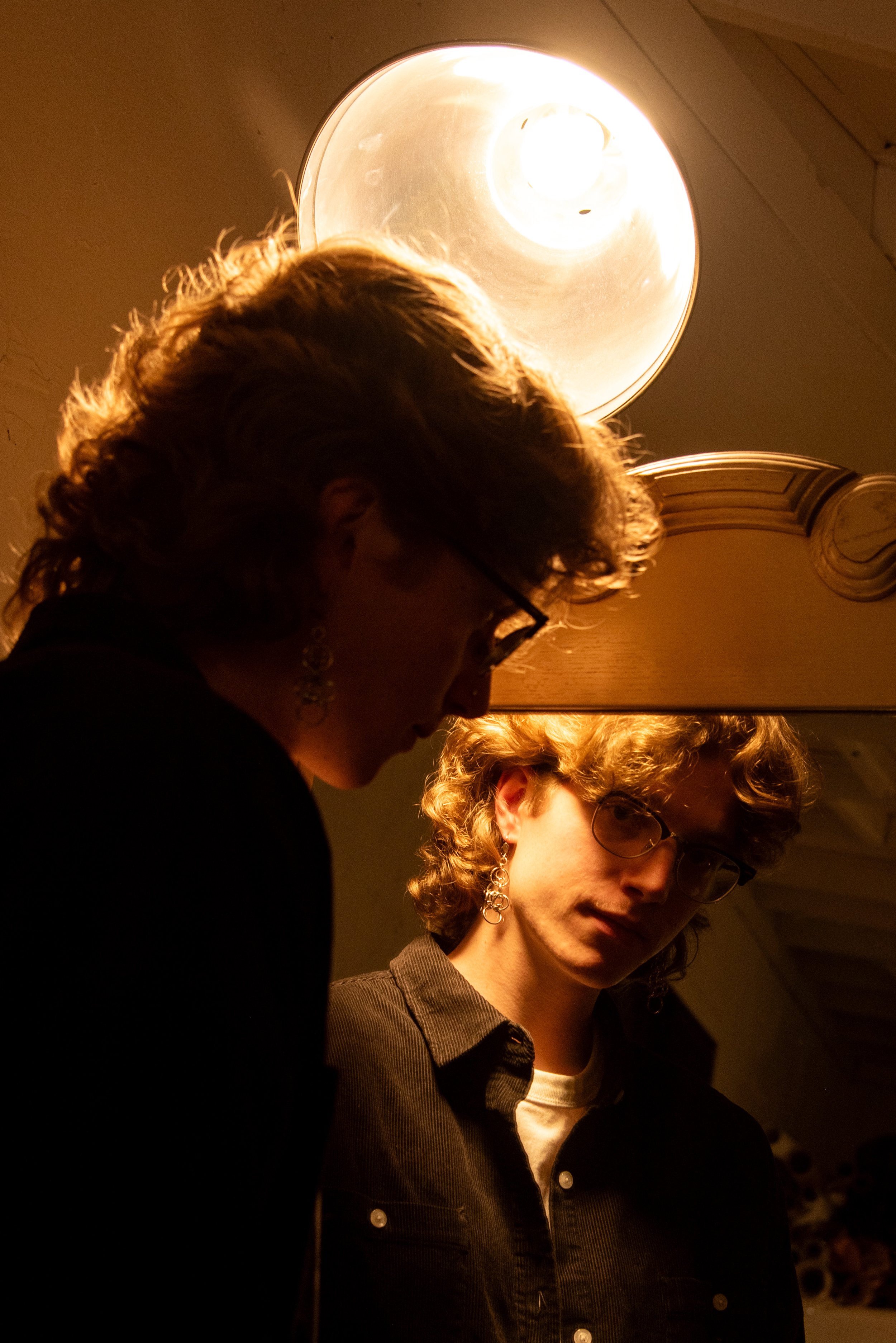 Designer Liam Roy looking at his reflection in a mirror in a dim lit, warm toned room,  wearing glasses and chainmail earrings and a corduroy buttoned shirt.