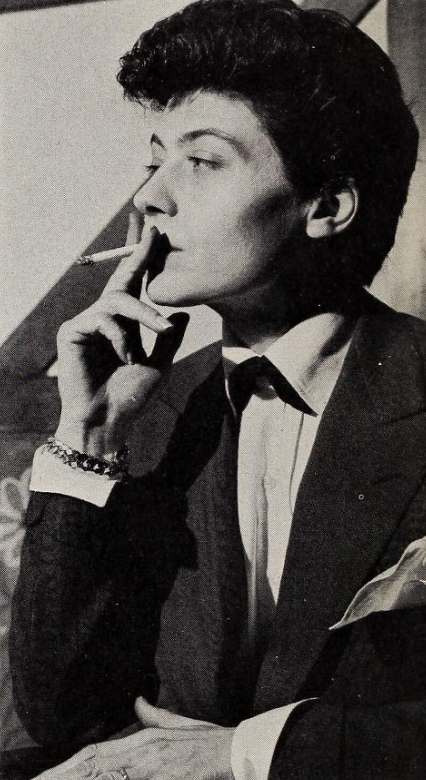 Profile view of Frede Baule smoking a cigarette, looking into the distance, and wearing a button up, blazer, chain bracelet, and ring on their ring finger.