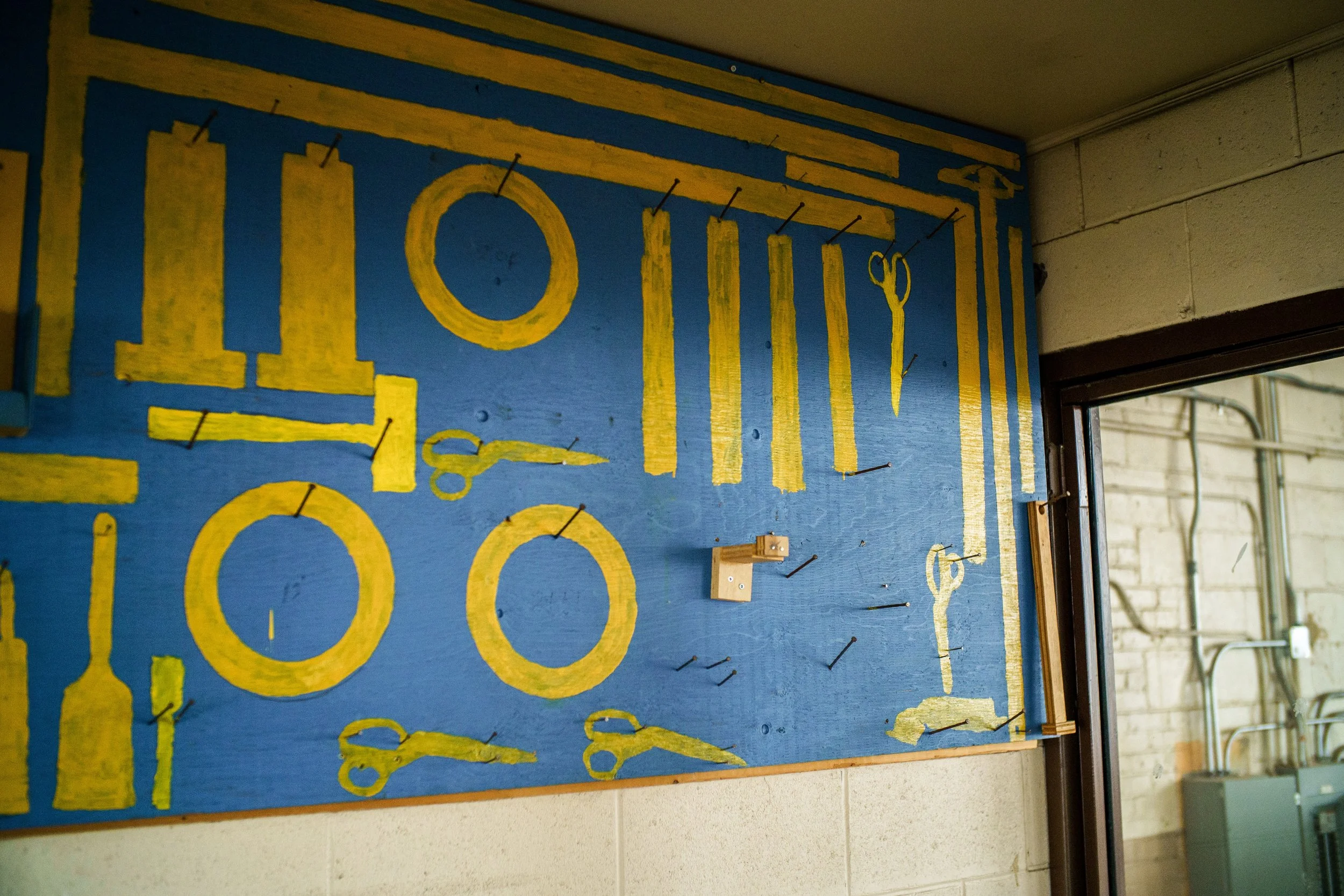 Photograph showing a blue wall with yellow outlines of various tools, including scissors, rolls, and bottles, arranged in an organized pattern. Empty hooks on the wall indicate where the actual tools are meant to be hung.