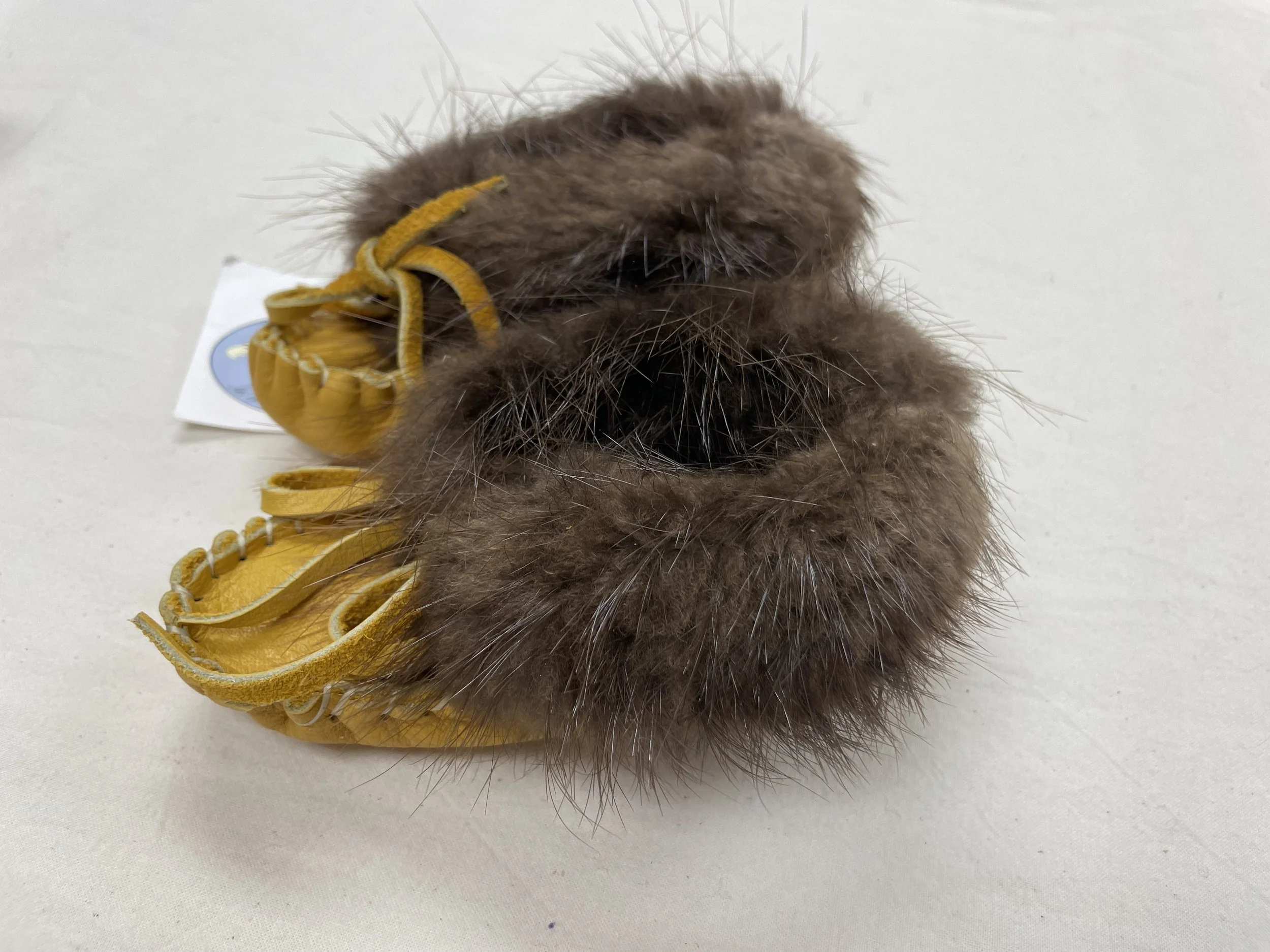 Side profile of a pair of small children's moccasins made from leather with brown fur trim around the ankles. Feature a tied lace on the front, placed on a light fabric surface with a tag partially visible.
