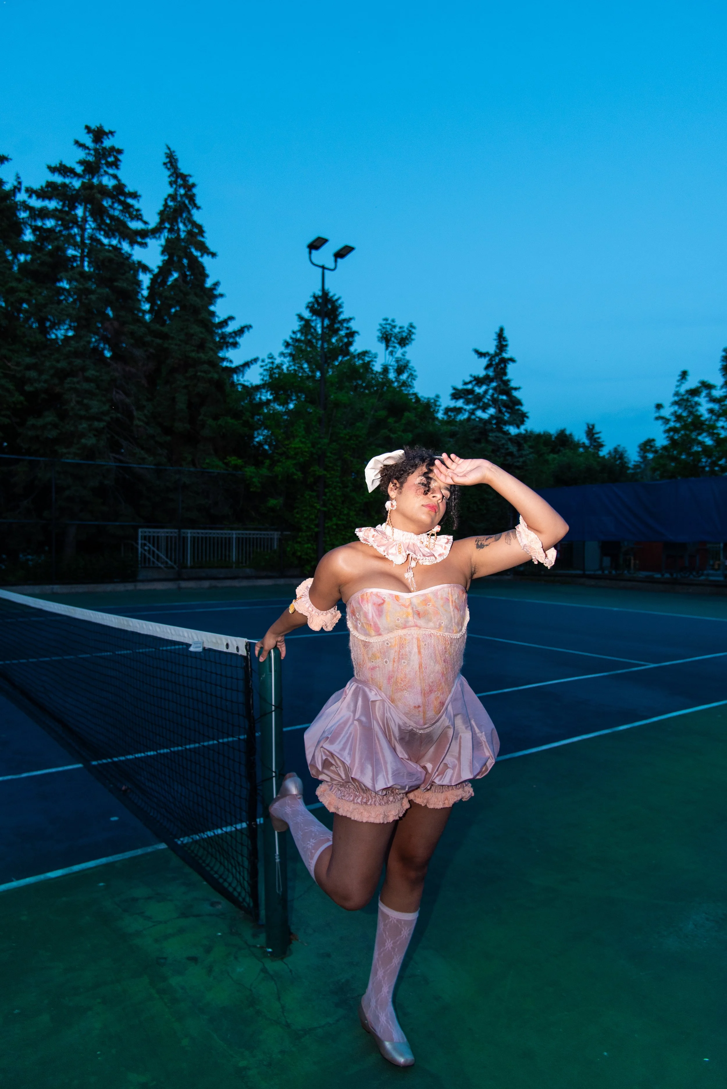 Designer Cami Leonelli Calzado posing on a tennis court at dusk, leaning on the net with one leg raised wearing a felted and silk baby pink and orange corset.