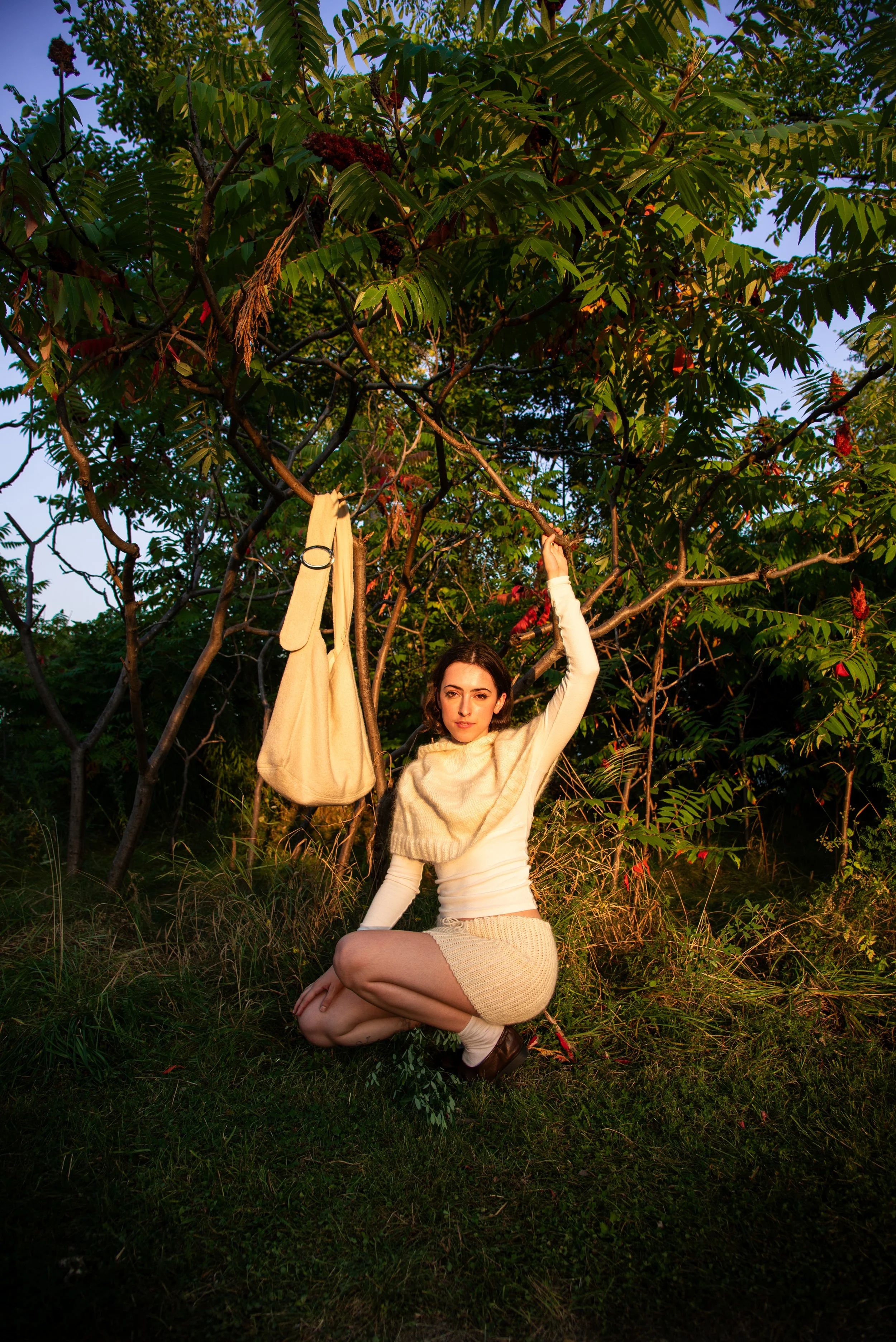 Designer Charlotte Little crouching beneath a leafy tree at golden hour, wearing a knitted cream shawl and skirt, with a knit cream bag hanging from a branch beside her.