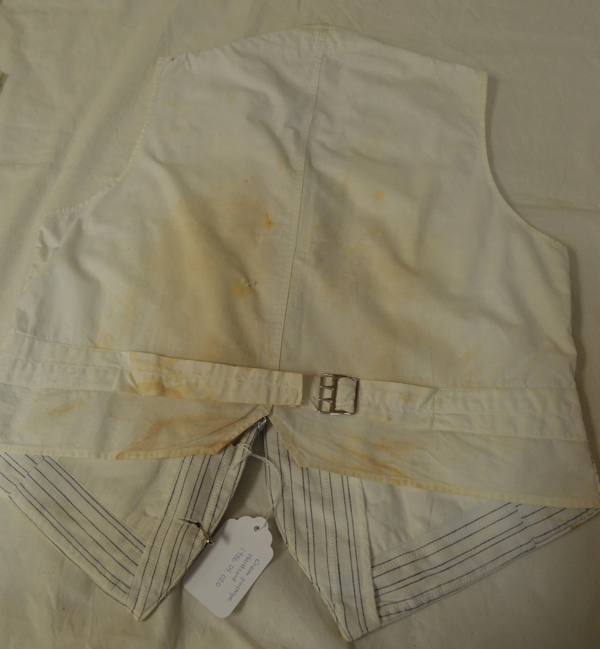 An external view of the back of the pinstripe vest. The back fabric is plain white cotton, as are the waist ties. The garment has heavy yellow and brown staining around the centre back and on the left tie.