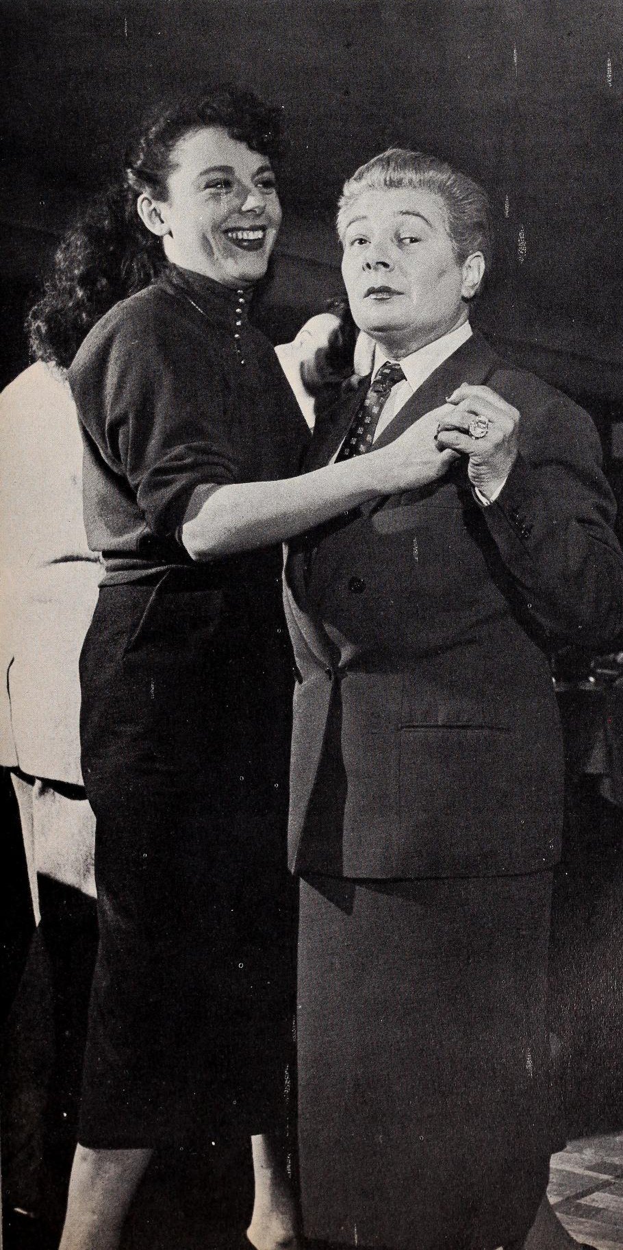 Moune wearing a pencil skirt and blazer with a patterned tie dancing hand in hand with a femme presenting figure at their bar Chez Moune.