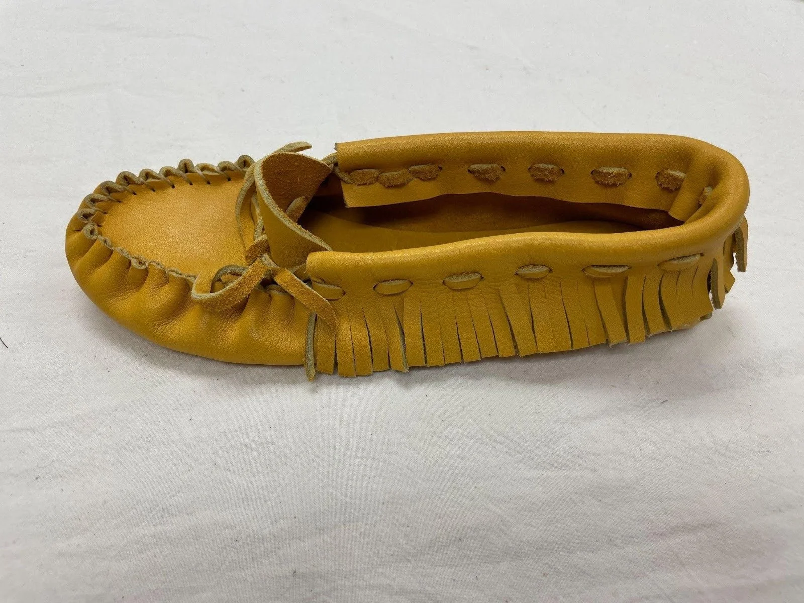 Side view of a single moccasin with leather fringe detailing along the sides and leather laces tied in bows on top on a white fabric surface.