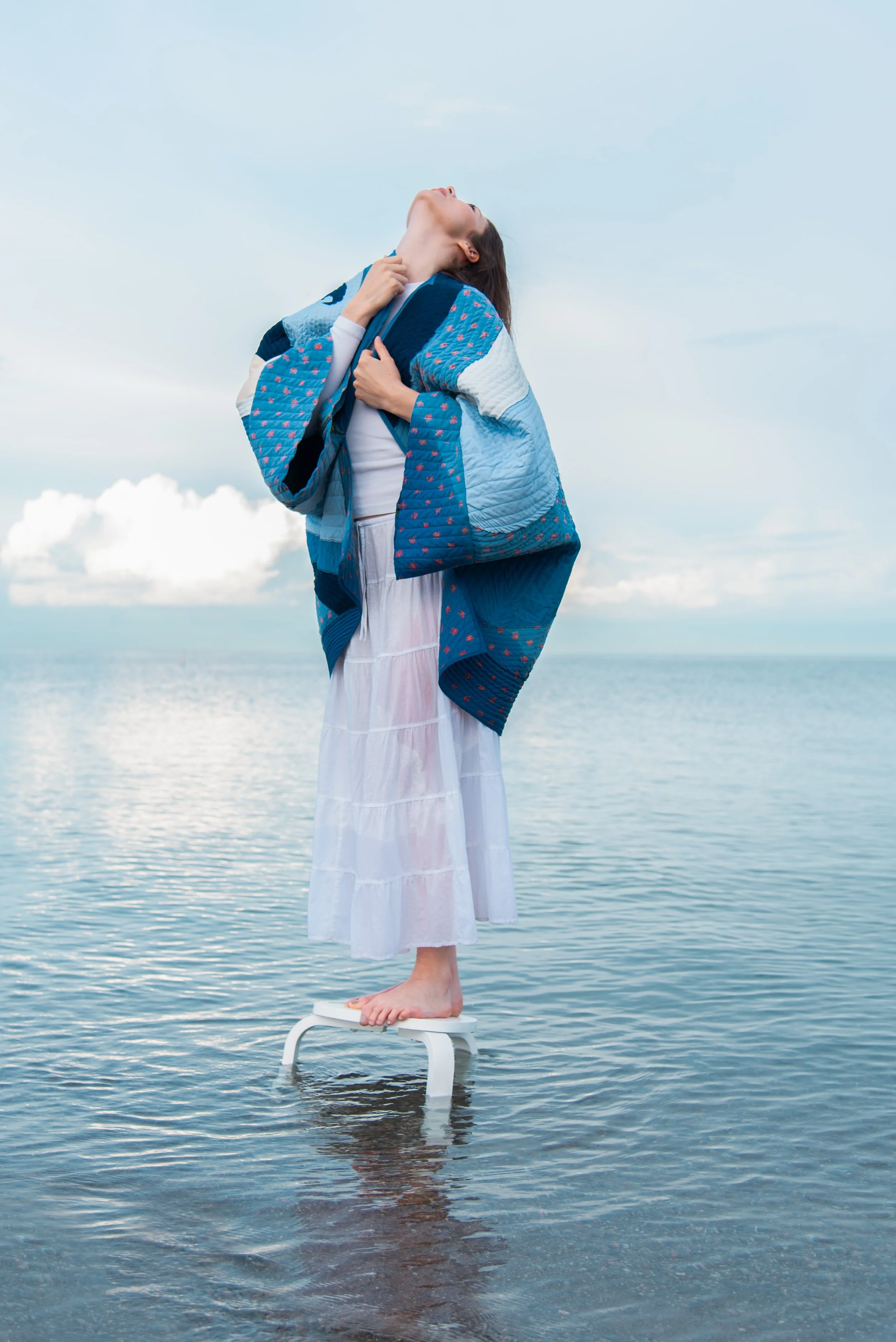 Designer Millie Yates standing on a stool in a large body of water surrounded by fluffy clouds looking up at the sky, she is wearing a blue and white quilted cotton jacket and a white skirt.