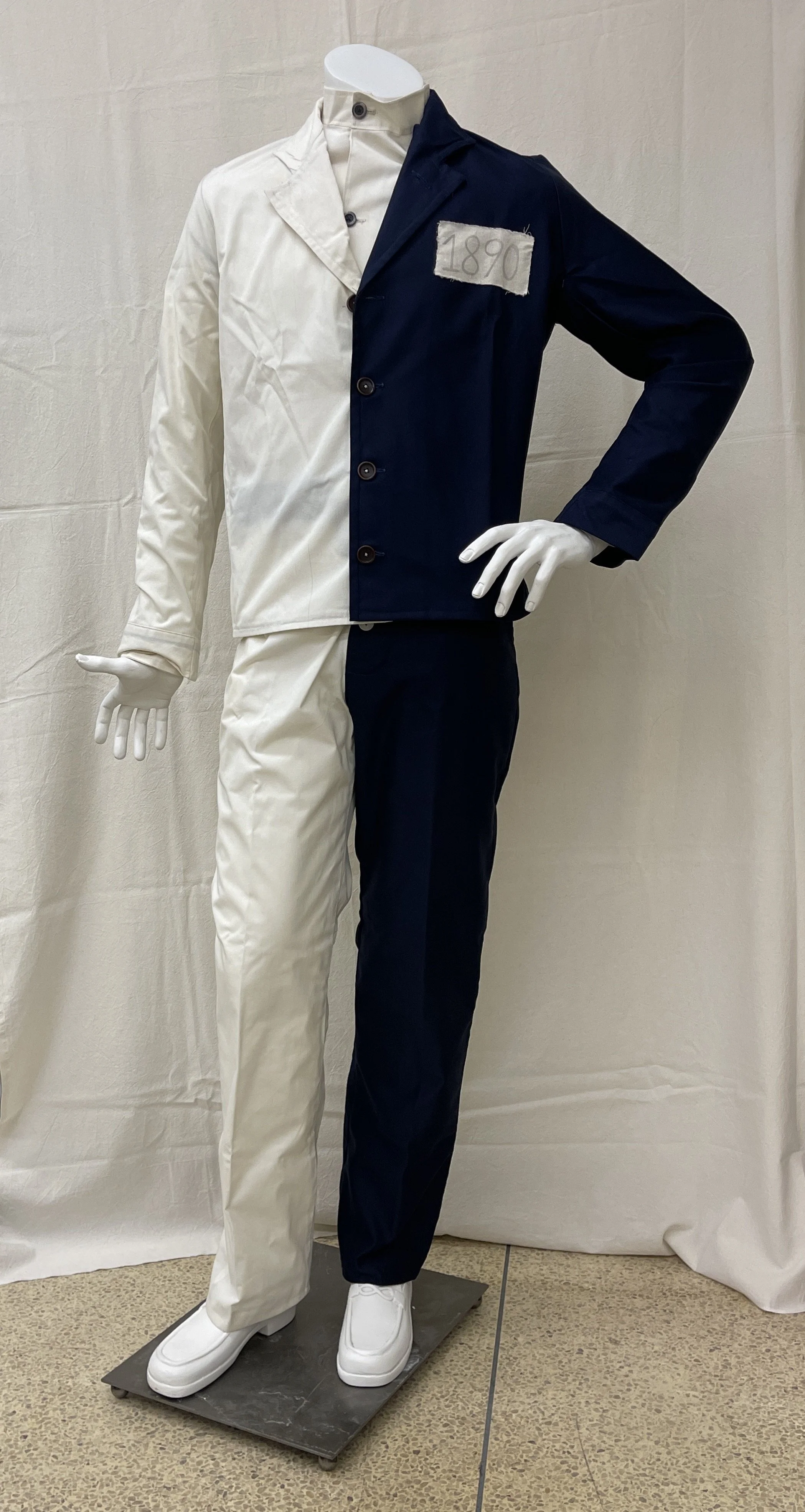 Photograph of a mannequin dressed in a half-black, half-white suit with matching pants, split vertically down the middle. The suit features a white patch with black numbers reading "1890" on the black side of the chest.