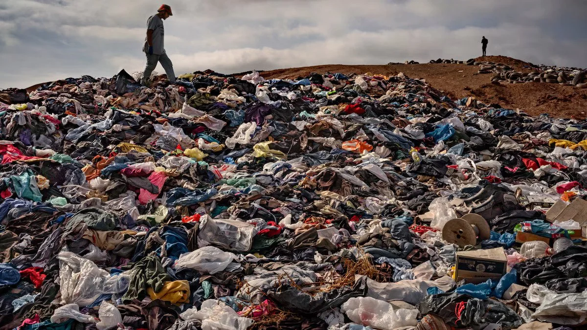 A person stands on a landfill of discarded clothes against a backdrop of cloudy sky. The silhouette of another person stands in the distance in the top right corner of the frame.