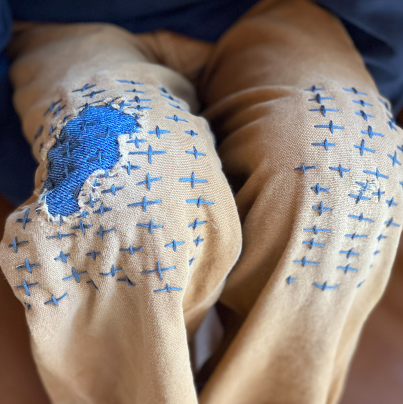 Sashiko mending with blue stitches and a blue fabric scrap on the knee of a pair of white jeans