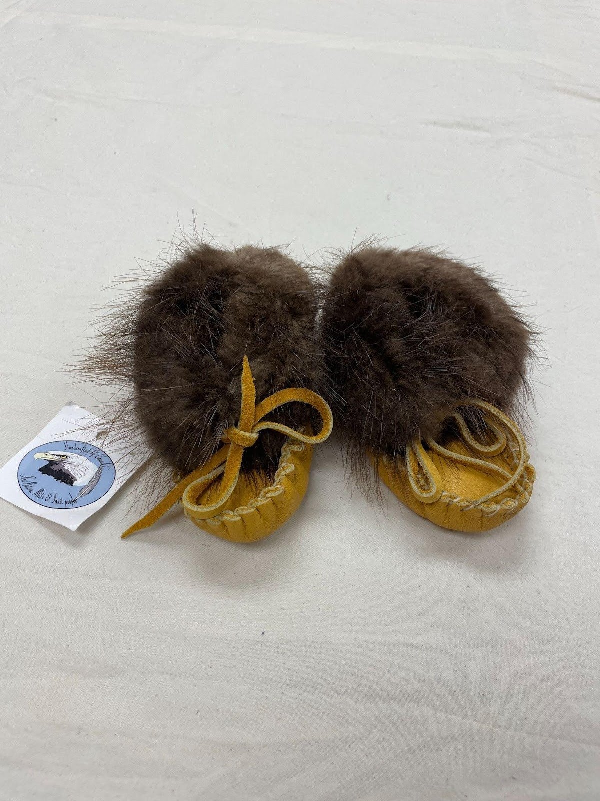 Pair of small children's moccasins with dark brown fur around the ankles, featuring leather laces tied in bows. A sales tag with an eagle logo and text is attached to one moccasin.