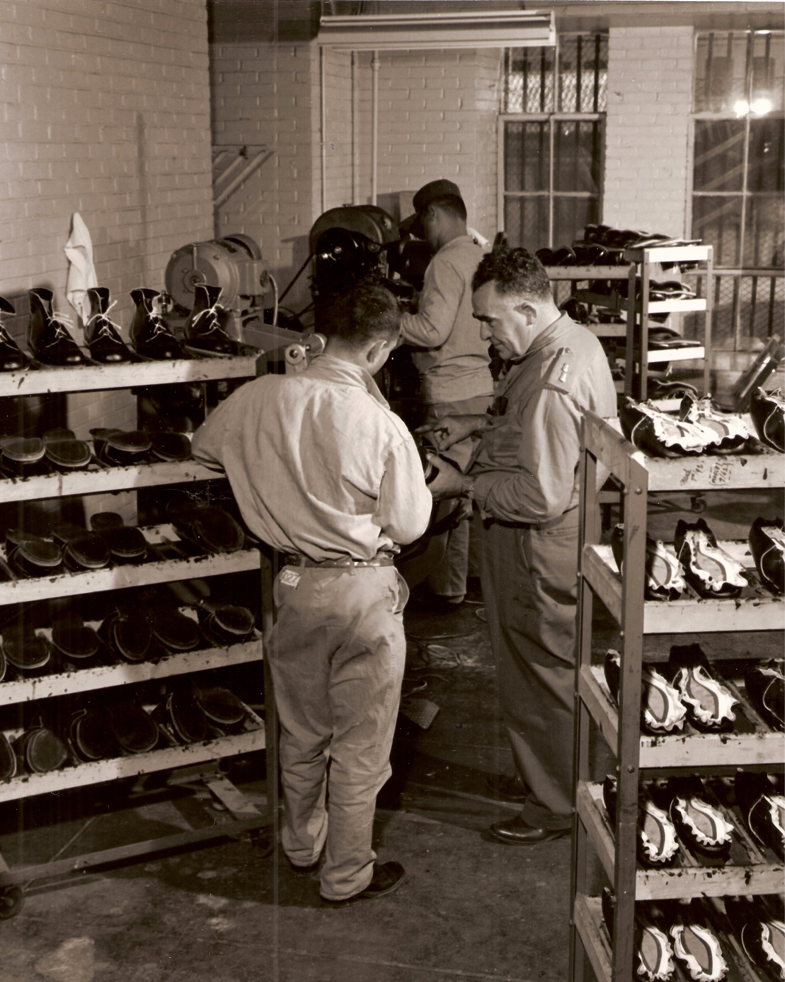 Three workers inspecting and organizing shoes in a factory or workshop setting. Multiple shelves filled with pairs of shoes surround the workers.