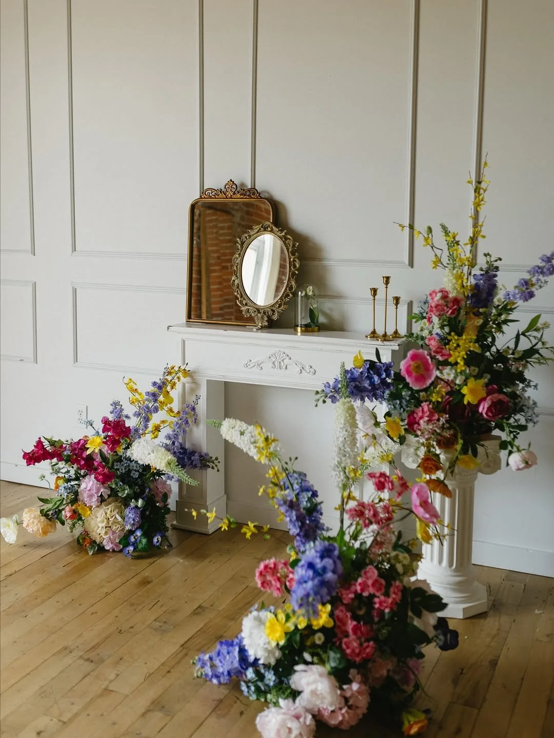 Decorative white fireplace mantle adorned with a vintage mirror, gold candlesticks, and small decorative items, with multiple large colorful flower arrangements on the floor and one on a pedestal.