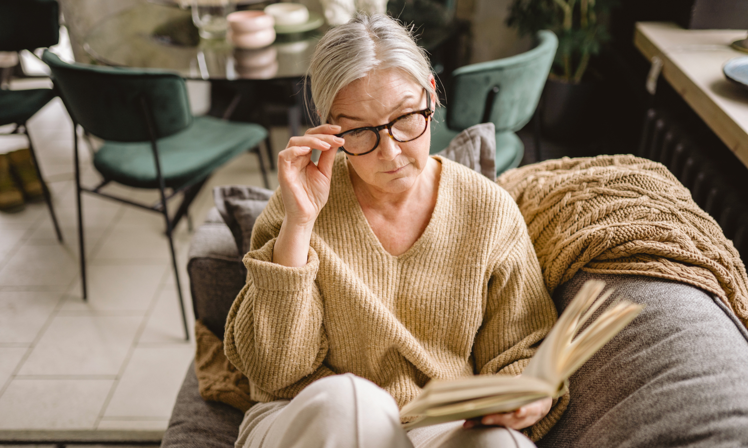 An older woman reading a book at home in a cozy sweater and glasses