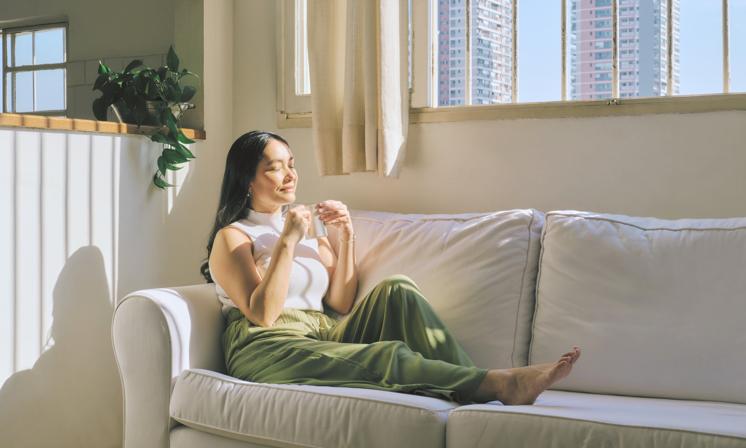 A woman looking content sitting on the coach with a tea and her eyes closed while the sun is on her face