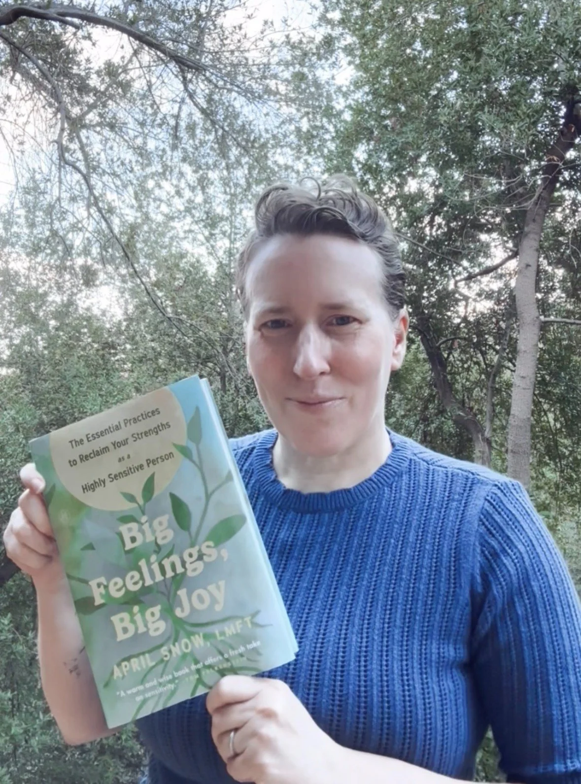 April Snow holding up a copy of her book Big Feelings, Big Joy, outside in front of trees